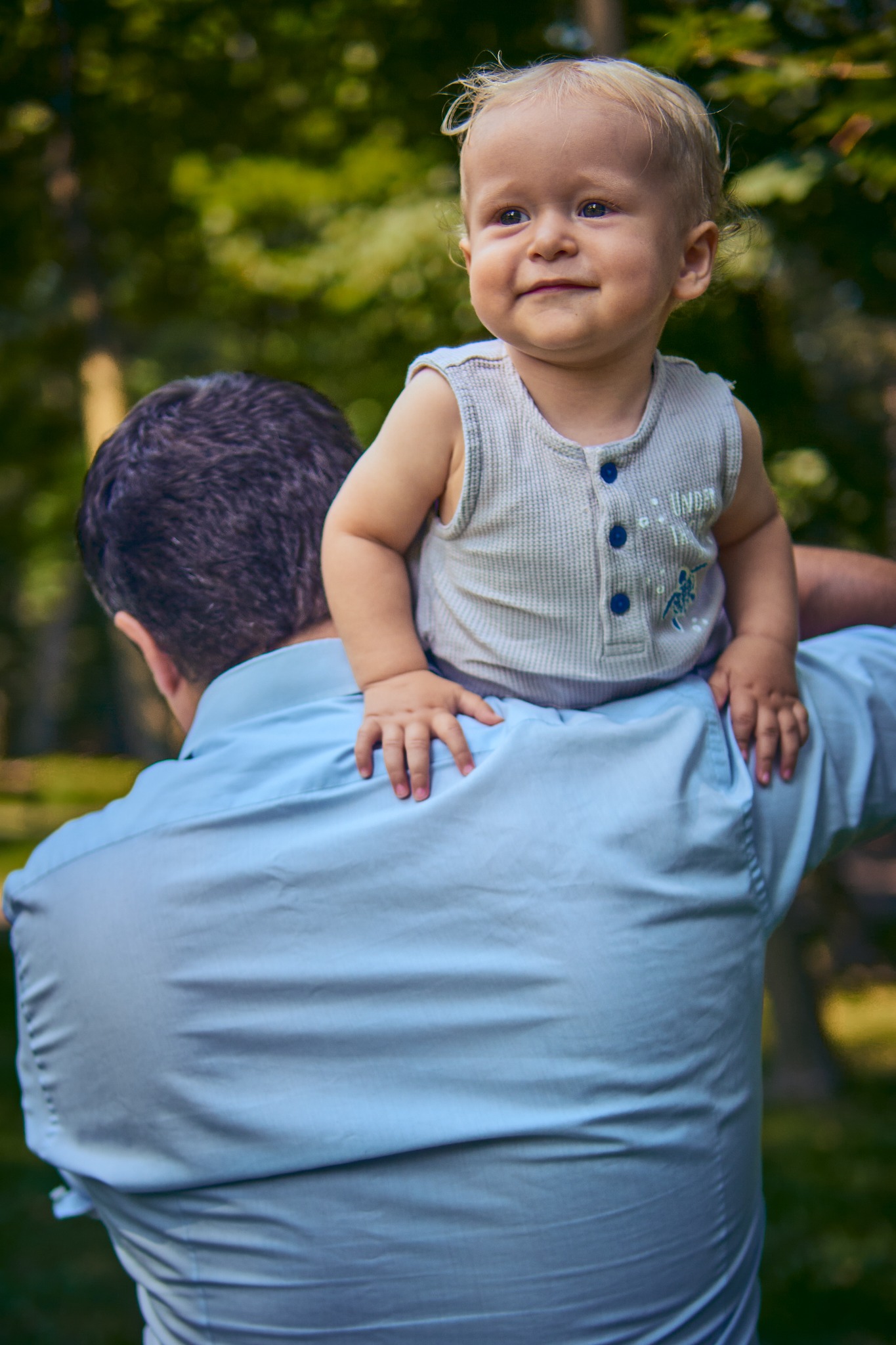 Uśmiechnięty roczny chłopiec z blond włosami siedzi na ramionach mężczyzny w błękitnej koszuli, na tle rozmytej zieleni.