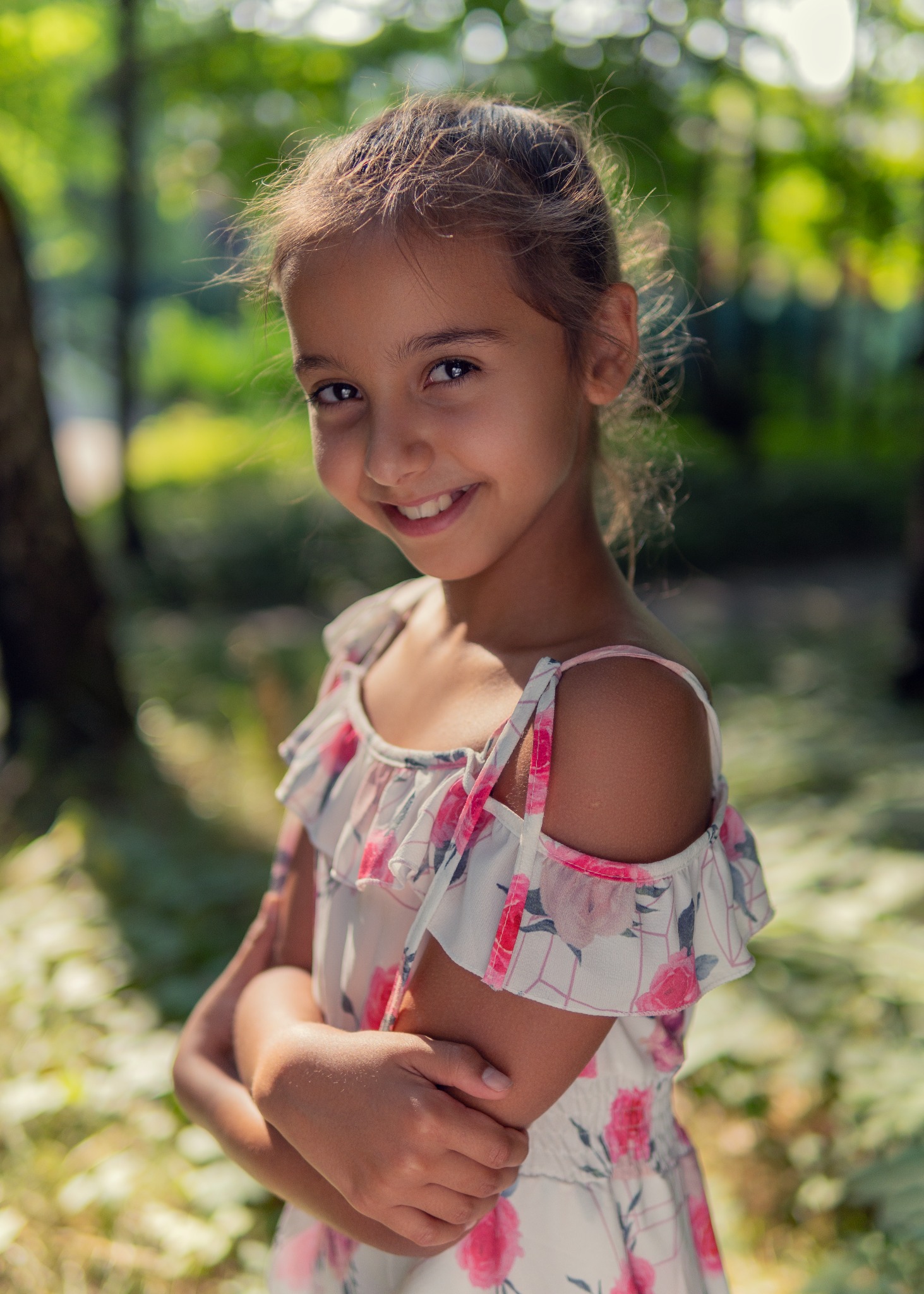 Portret uśmiechniętej dziewczynki w letniej sukience w kwiaty, stojącej na tle zamazanej zieleni parku.