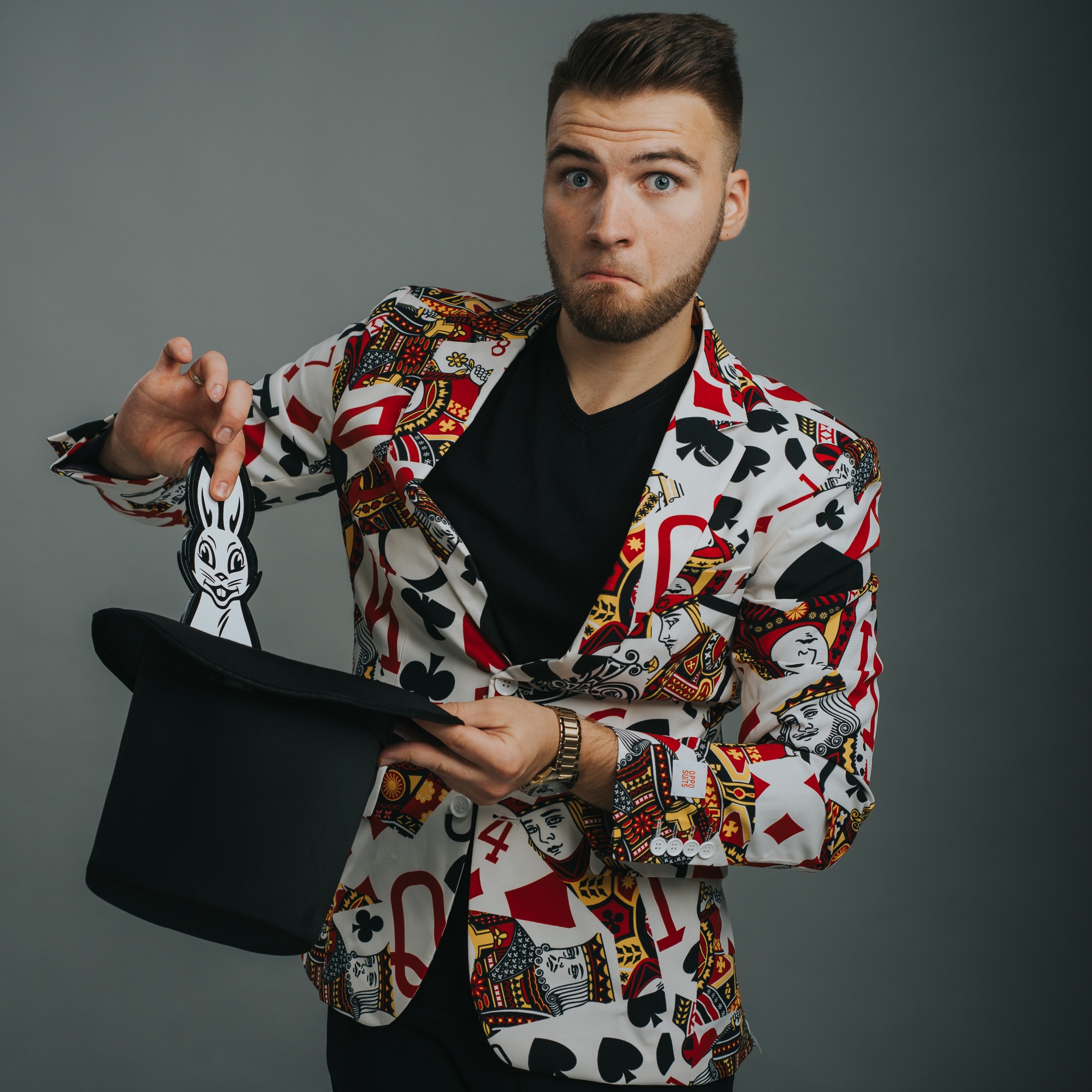 Mężczyzna w marynarce w karty trzyma czarny cylinder i kartonowego królika, patrząc z zaskoczeniem.