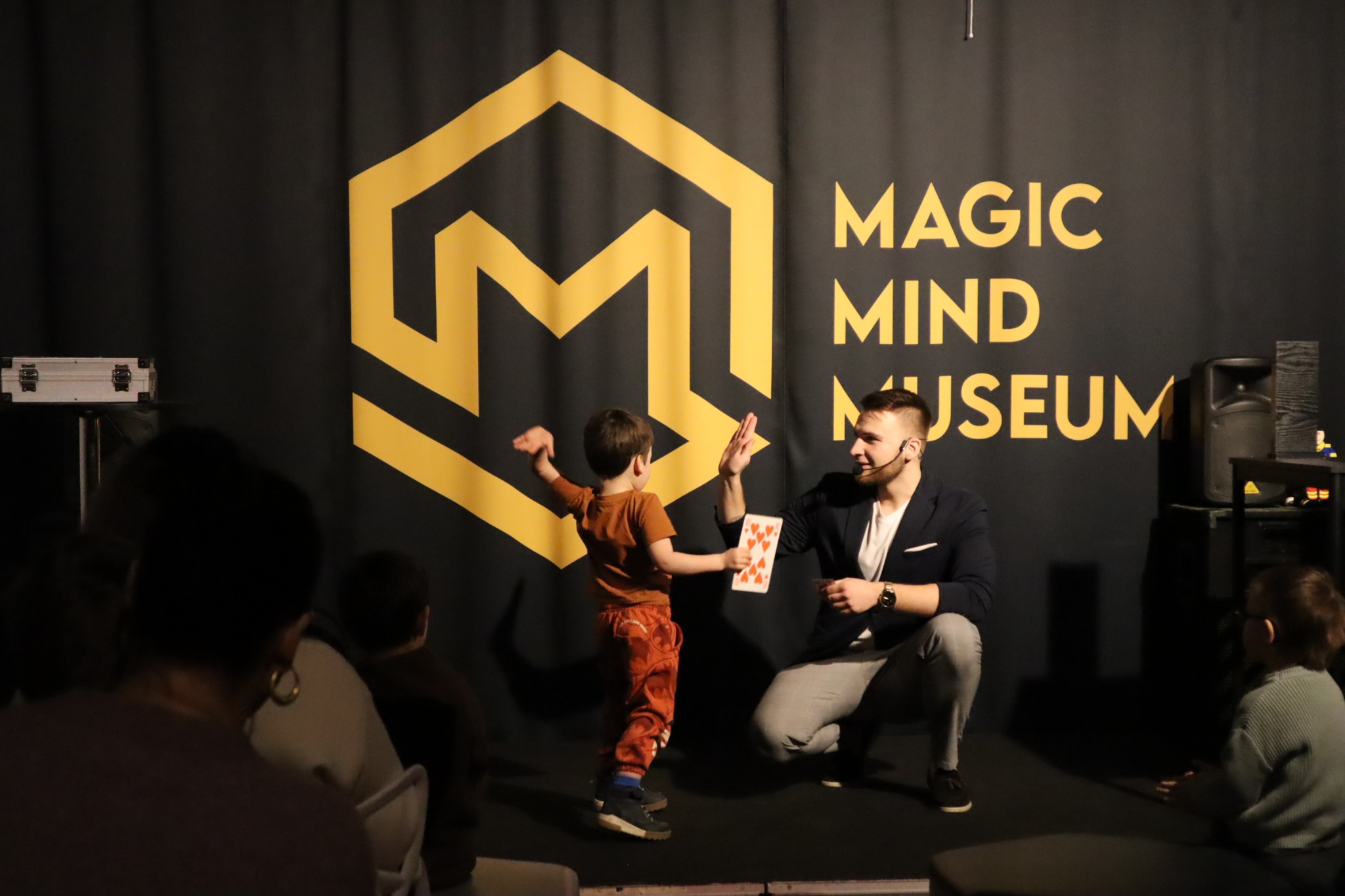 Iluzjonista w ciemnej marynarce prezentuje kartę z czerwonymi sercami chłopcu podczas występu w Magic Mind Museum, na czarnym tle z żółtym logo i napisem.