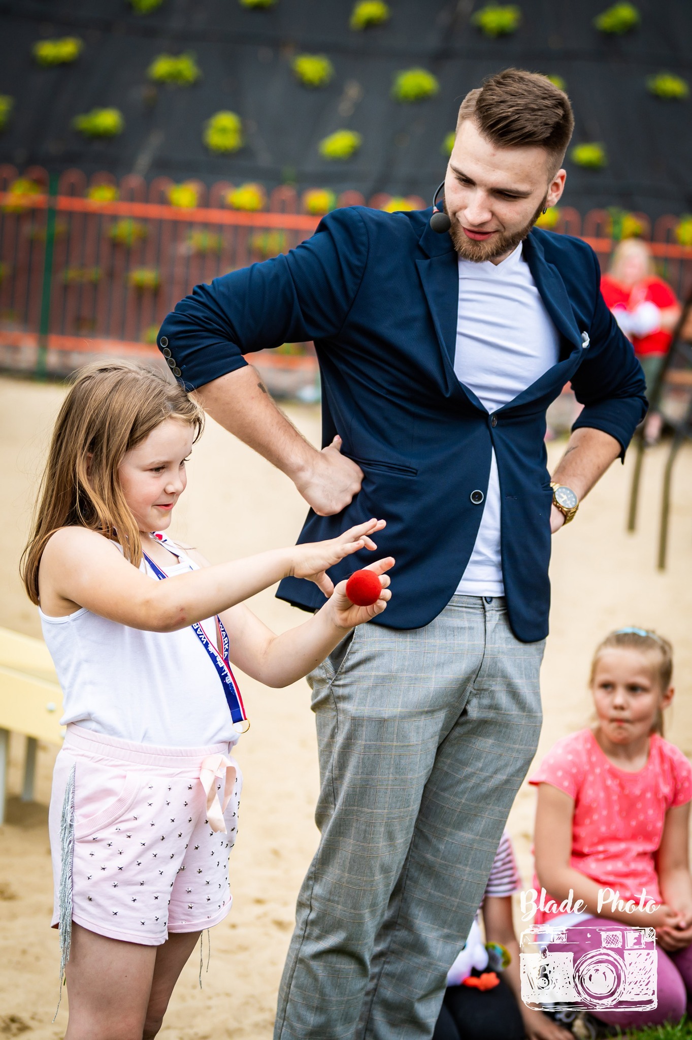 Iluzjonista w granatowej marynarce i szarych spodniach prezentuje trik z czerwoną kulką dziewczynce w białej koszulce i różowych spodenkach z frędzlami, na tle plenerowej sceny.