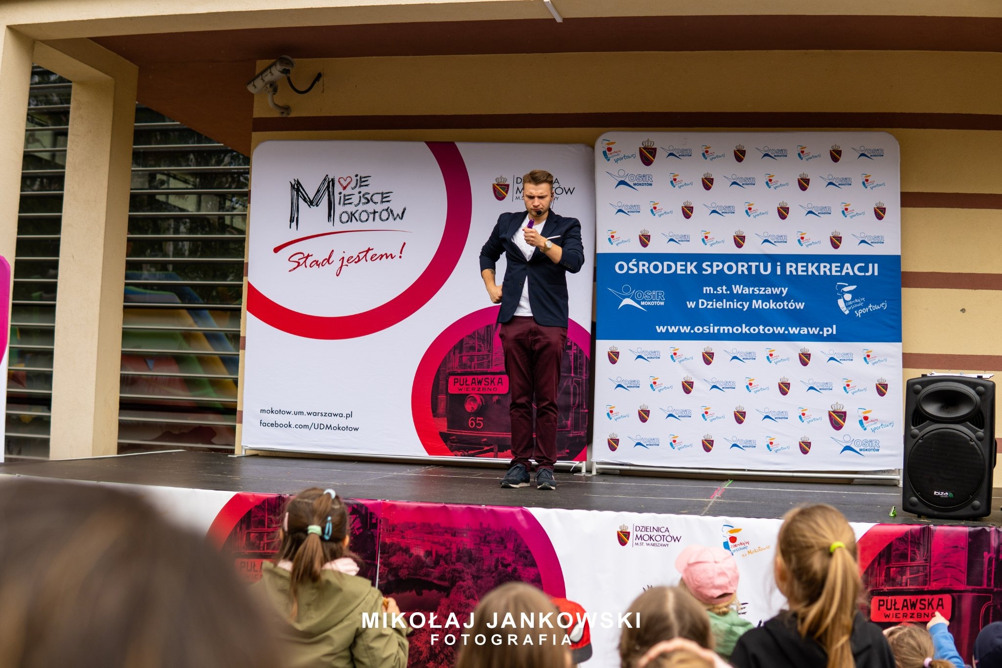 Mężczyzna w marynarce na scenie podczas wydarzenia plenerowego, trzymający mikrofon, na tle banerów z logo dzielnicy Mokotów i hasłem 'Stąd jestem!', widownia dziecięca w pierwszym planie.