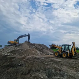 Żółta koparka Volvo na szczycie hałdy gruzu i ziemi, obok żółto-czarna koparko-ładowarka JCB, na pierwszym planie ślady opon na błotnistej ziemi, pochmurne niebo w tle.