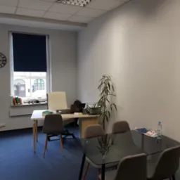 Jasne biuro z biurkiem, krzesłami, rośliną doniczkową i szklanym stołem konferencyjnym z butelką wody i magazynem, widok z perspektywy gościa.