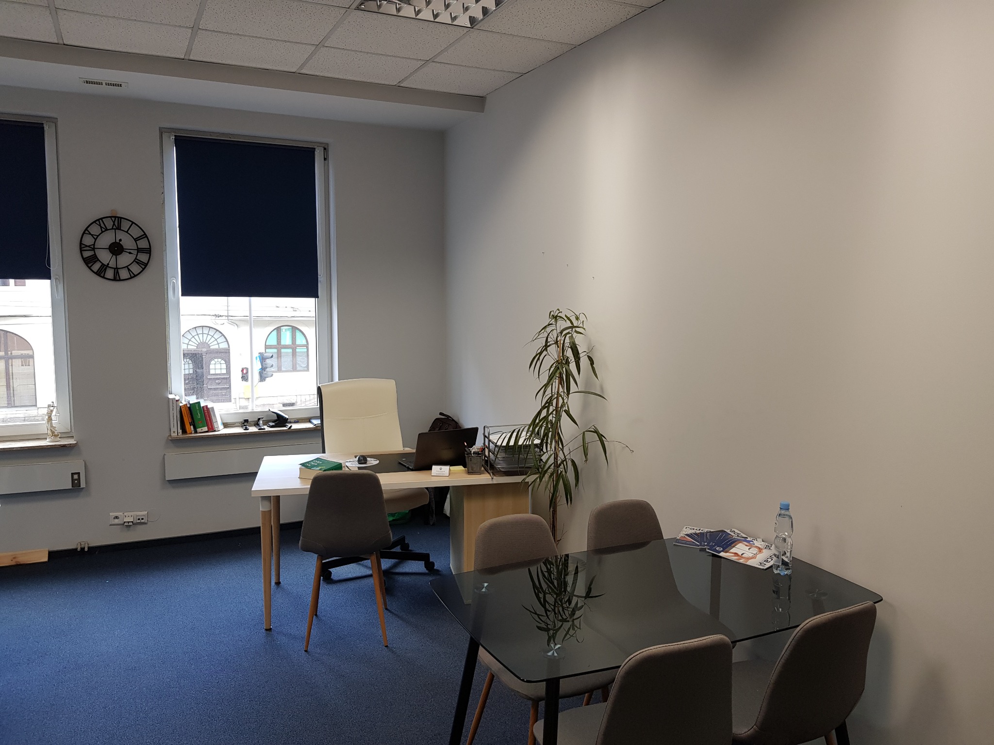 Jasne biuro z biurkiem, krzesłami, rośliną doniczkową i szklanym stołem konferencyjnym z butelką wody i magazynem, widok z perspektywy gościa.