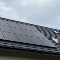 Czarny panel słoneczny na ciemnym dachu z dachówki, widok z dołu pod kątem, widoczne okna dachowe i komin, pochmurne niebo.