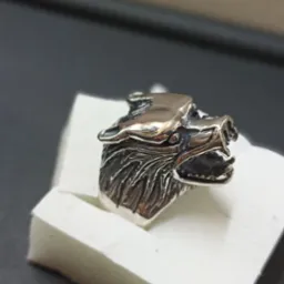 Srebrny pierścień w kształcie wilczej głowy z wyraźnymi detalami sierści, umieszczony na białym pudełku na tle czarnej powierzchni.