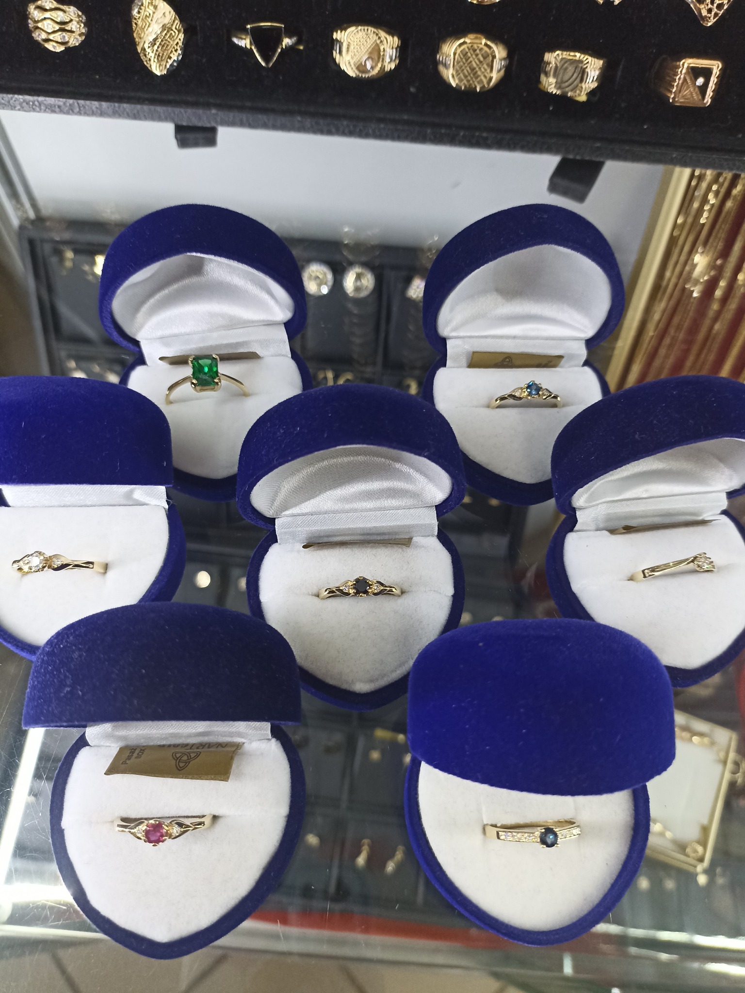 Siedem złotych pierścionków z różnymi kamieniami szlachetnymi w granatowych, aksamitnych pudełkach, ustawionych na szklanej powierzchni ekspozycyjnej, z widocznymi innymi pierścionkami i złotymi...