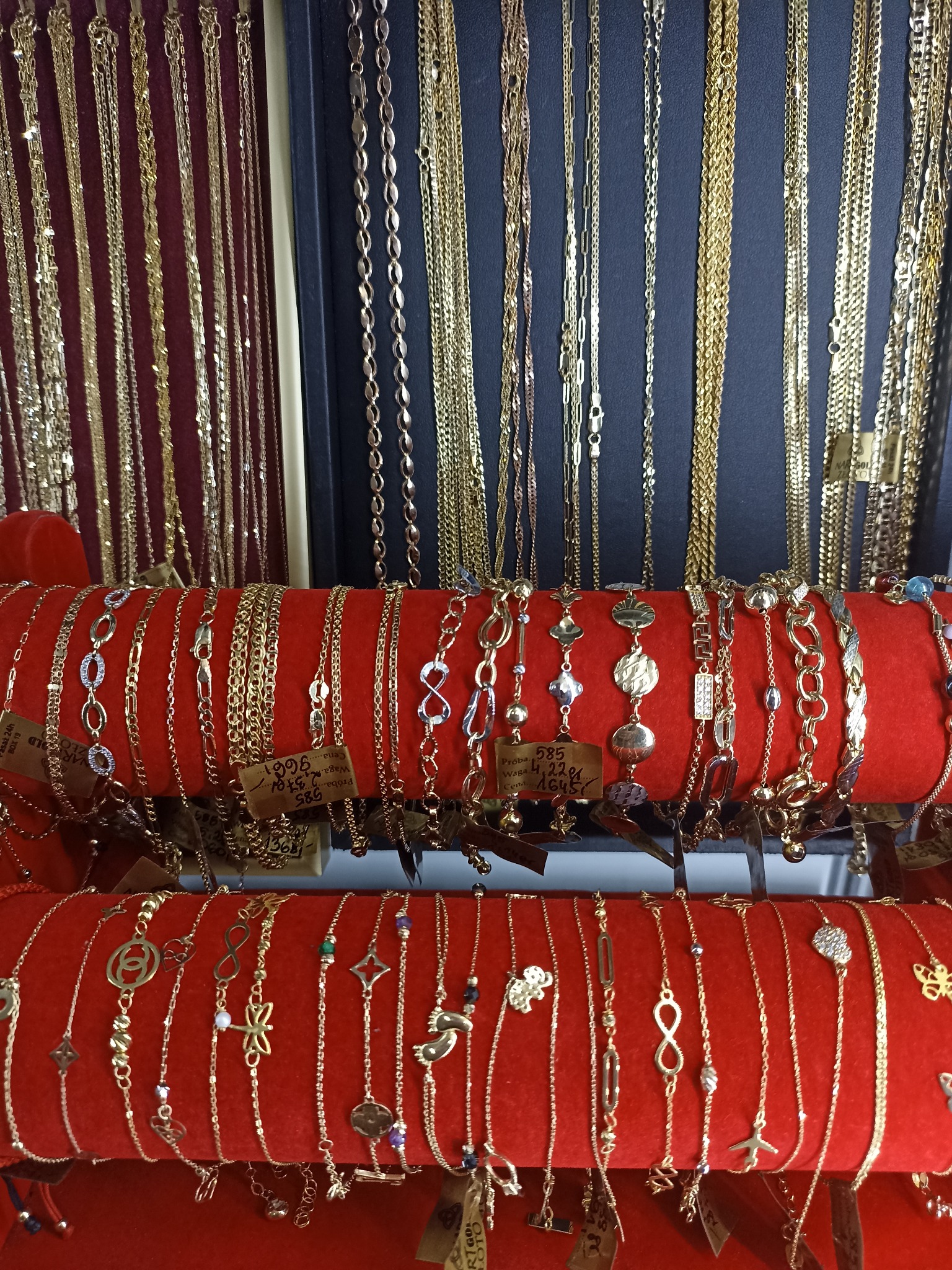 Ekspozycja złotej biżuterii: naszyjniki i bransoletki na czerwonych stojakach, z widocznymi metkami cenowymi i próbami złota.