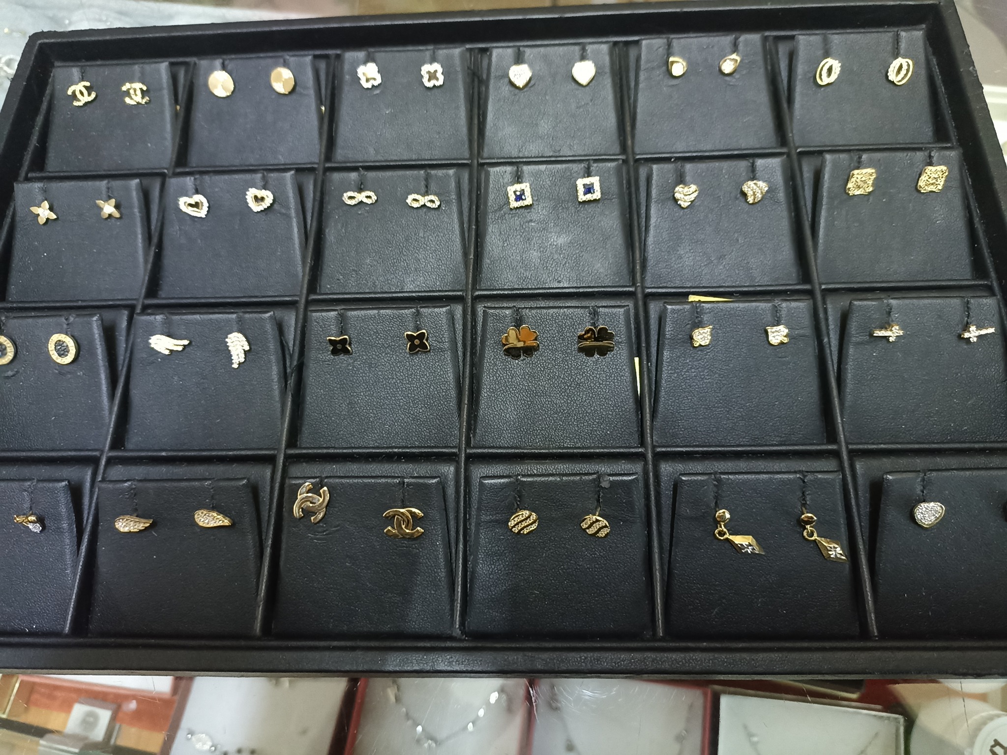 Ekspozycja różnorodnych złotych kolczyków na czarnym, podzielonym na przegródki ekspozytorze jubilerskim, prezentująca wzory od serc i koniczynek po logo Chanel i abstrakcyjne formy.