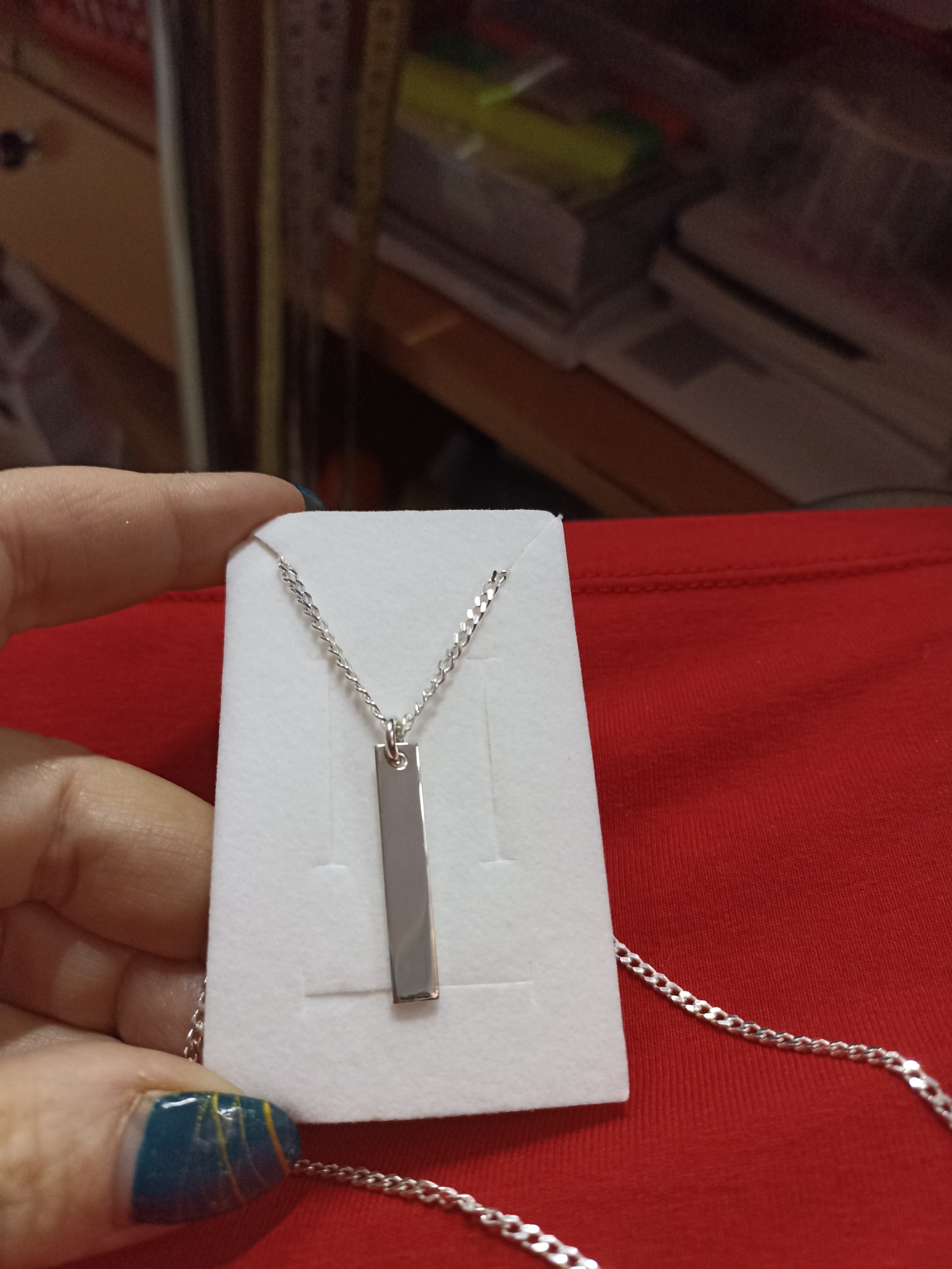 Srebrny naszyjnik z prostokątną zawieszką, prezentowany na białym kartoniku, trzymany w dłoni z niebiesko-złotym manicure na czerwonym tle.