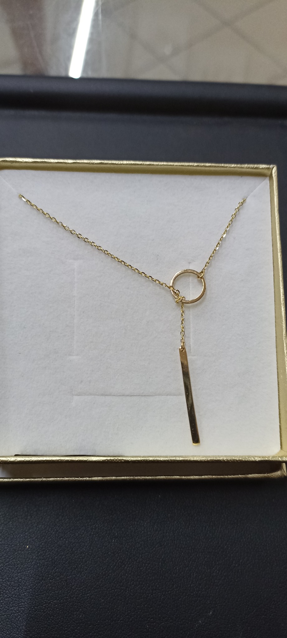 Delikatny złoty łańcuszek z okrągłym elementem i prostokątną zawieszką, umieszczony w eleganckim pudełku prezentowym z jasnym wnętrzem.