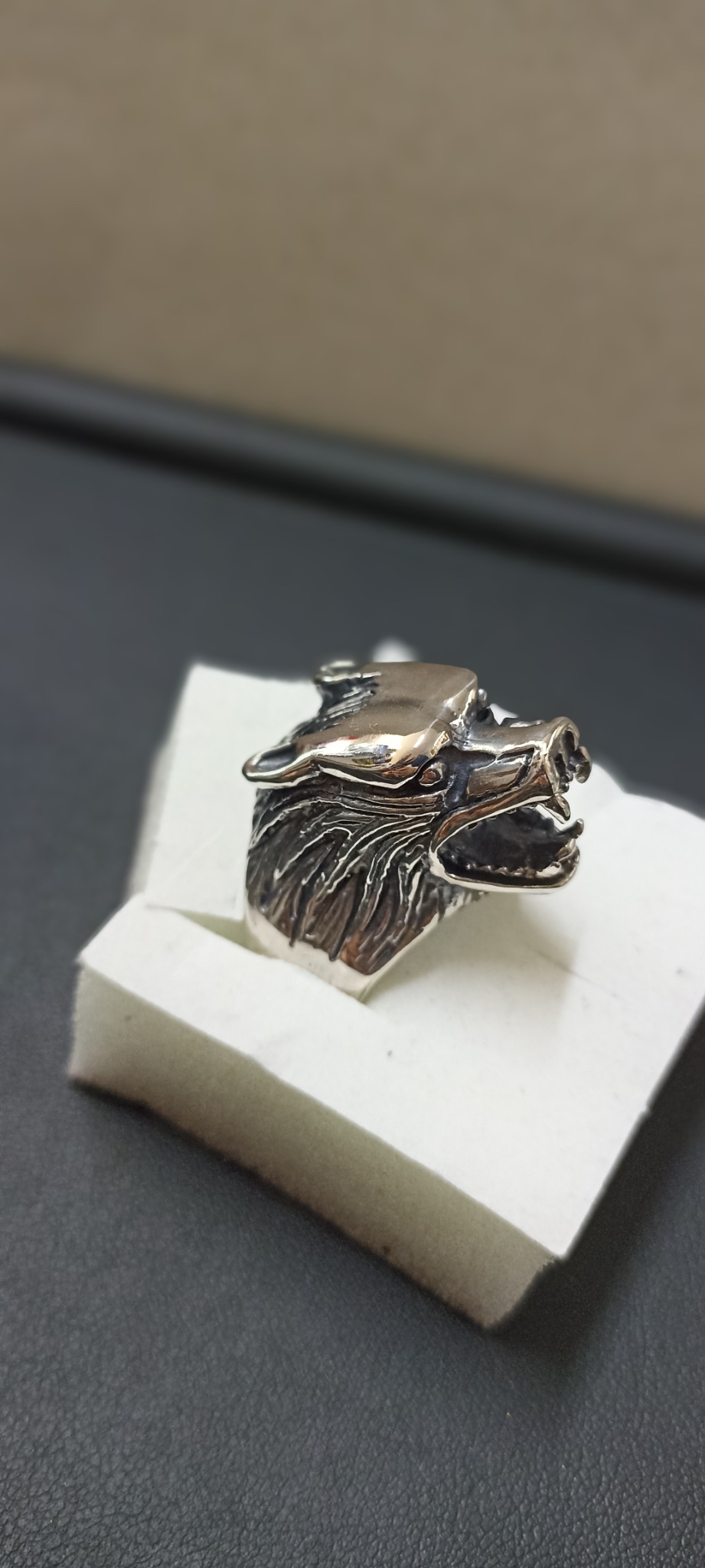 Srebrny pierścień w kształcie wilczej głowy z wyraźnymi detalami sierści, umieszczony na białym pudełku na tle czarnej powierzchni.