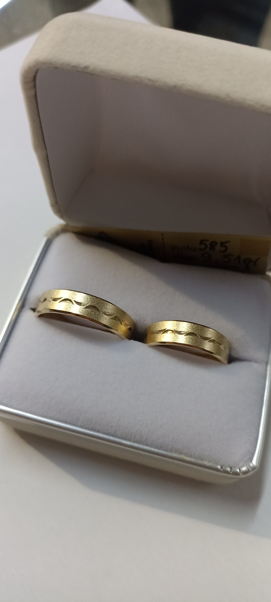 Dwa złote, matowe obrączki z delikatnym wzorem fali w welurowym pudełku, widoczny fragment metki z próbą złota 585.