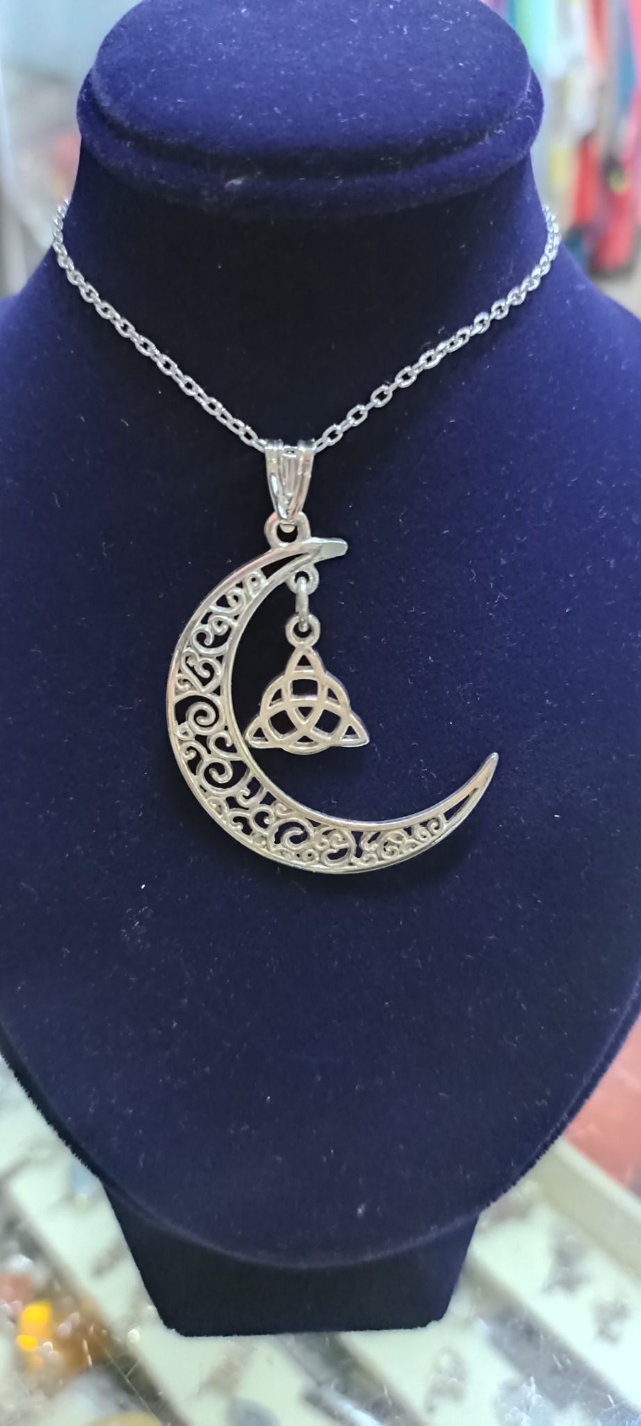 Srebrny wisiorek w kształcie księżyca z ażurowym wzorem i symbolem trójkąta celtyckiego zawieszony na łańcuszku, prezentowany na aksamitnym popiersiu.