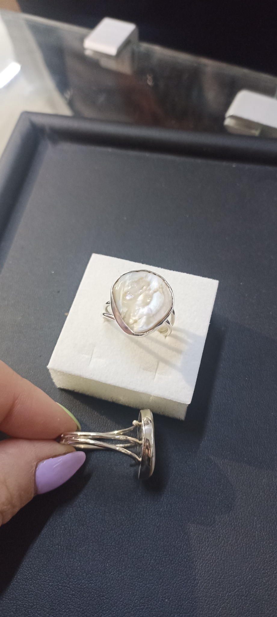 Dwa pierścionki z perłą w srebrnej oprawie, jeden na białym pudełku, drugi trzymany w dłoni z fioletowym lakierem do paznokci, na ciemnym tle.