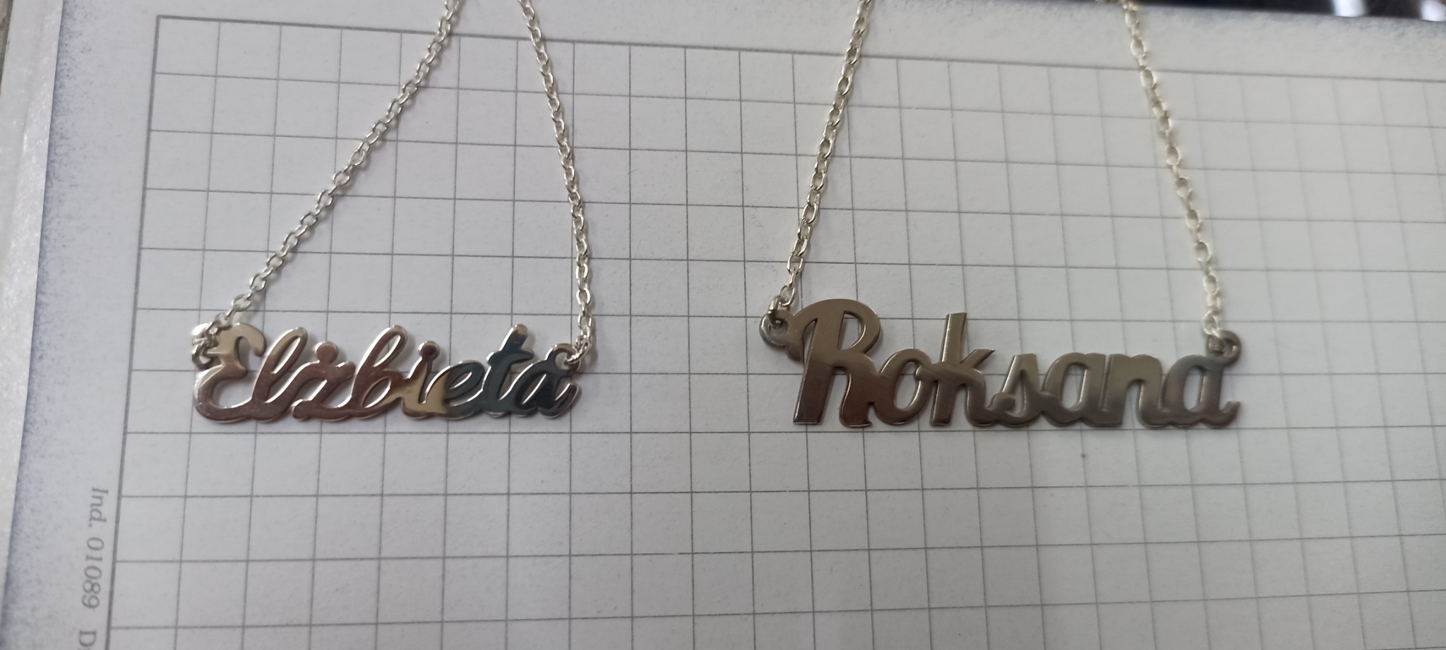 Dwa srebrne naszyjniki z imionami 'Elżbieta' i 'Roksana' leżą na kartce w kratkę.