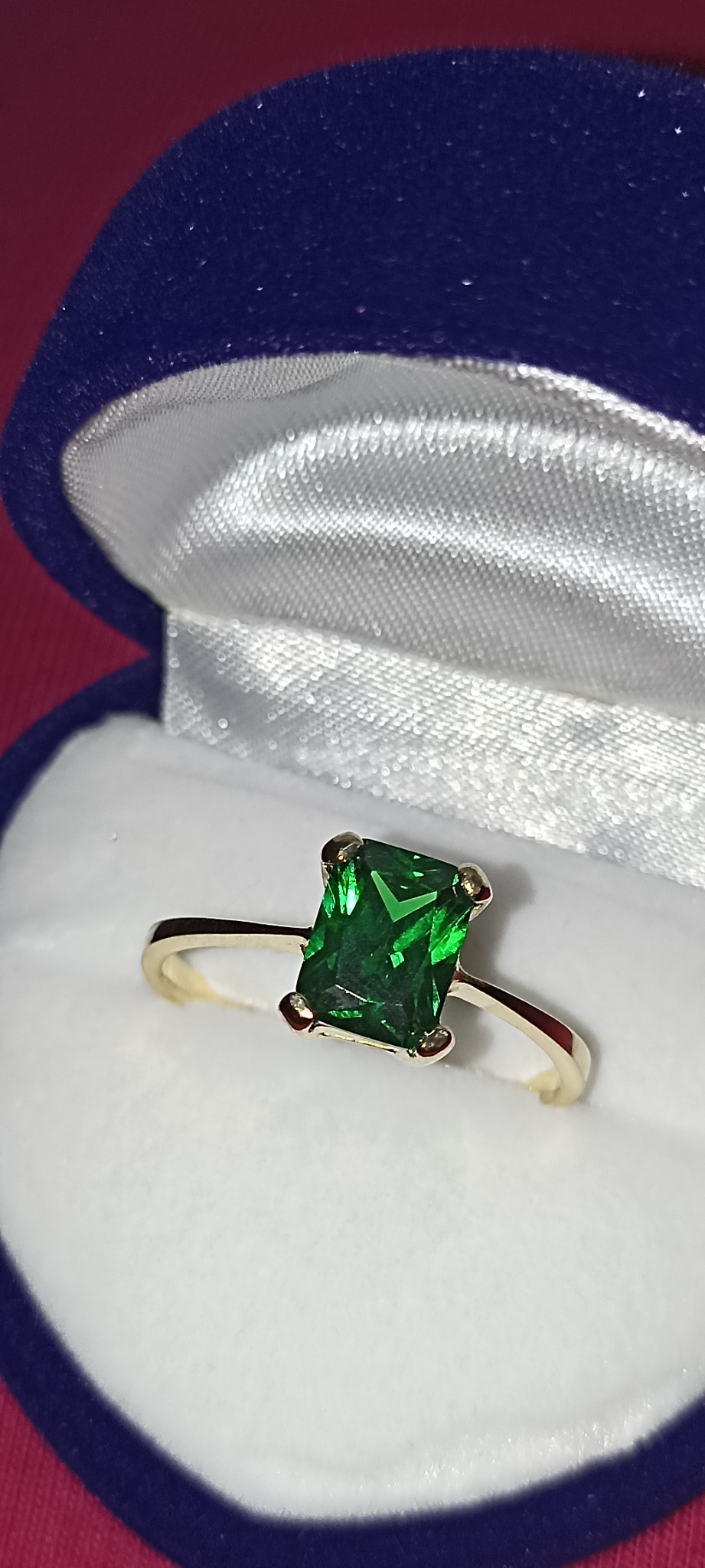 Pierścionek z prostokątnym szmaragdem na złotym pasku, prezentowany w niebieskim pudełku z satynową wyściółką.