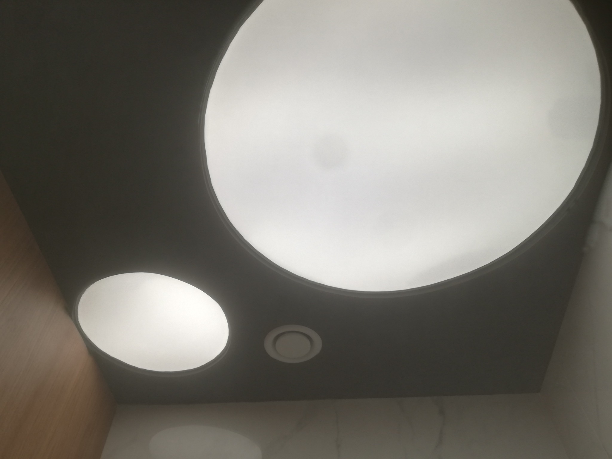 Nowoczesny sufit podwieszany z okrągłymi lampami LED, szara zabudowa, widoczny nawiewnik wentylacyjny, marmurowe wykończenie ściany.