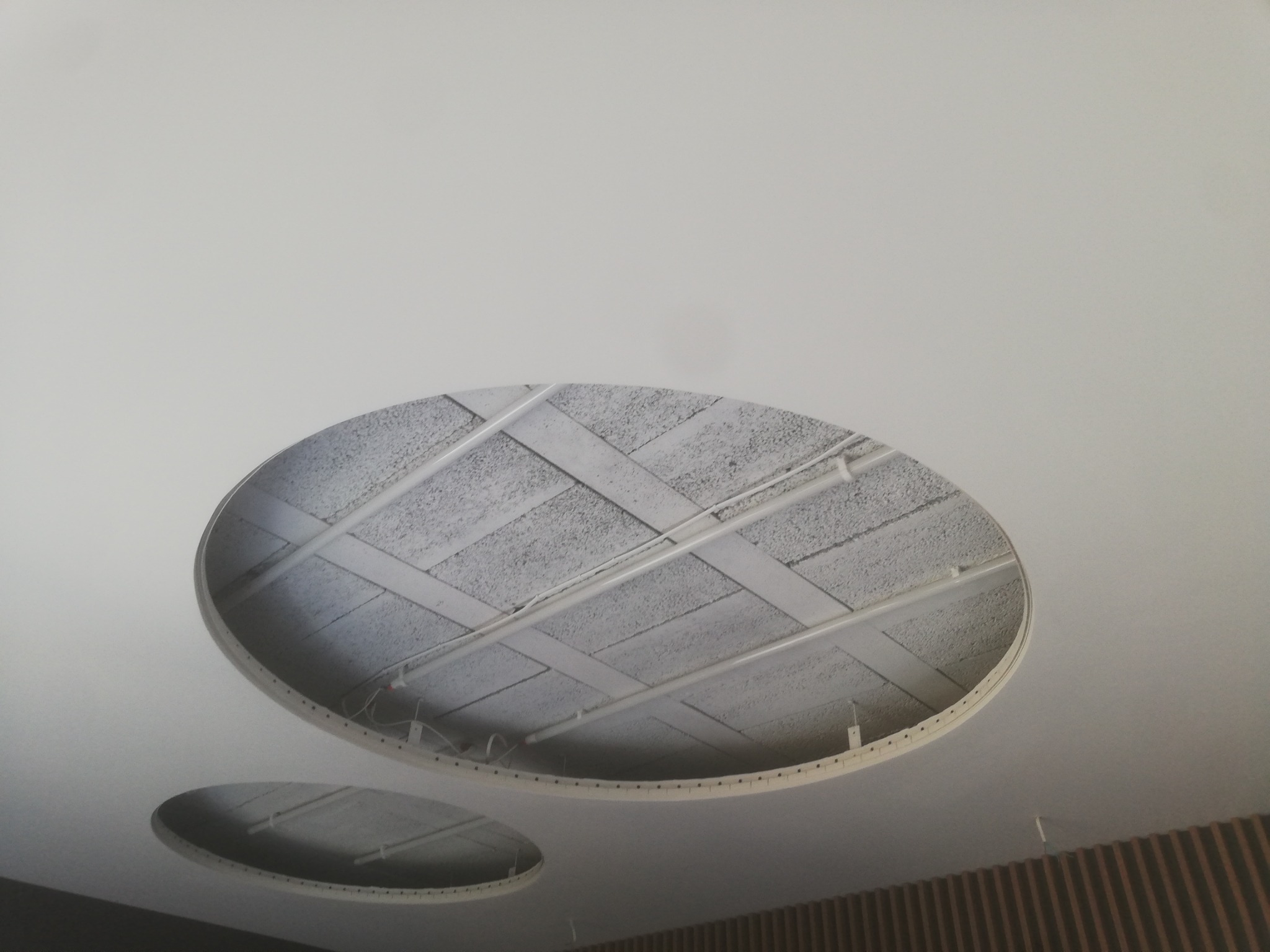 Owalne otwory w białym suficie podwieszanym, widoczne elementy konstrukcyjne i instalacje.