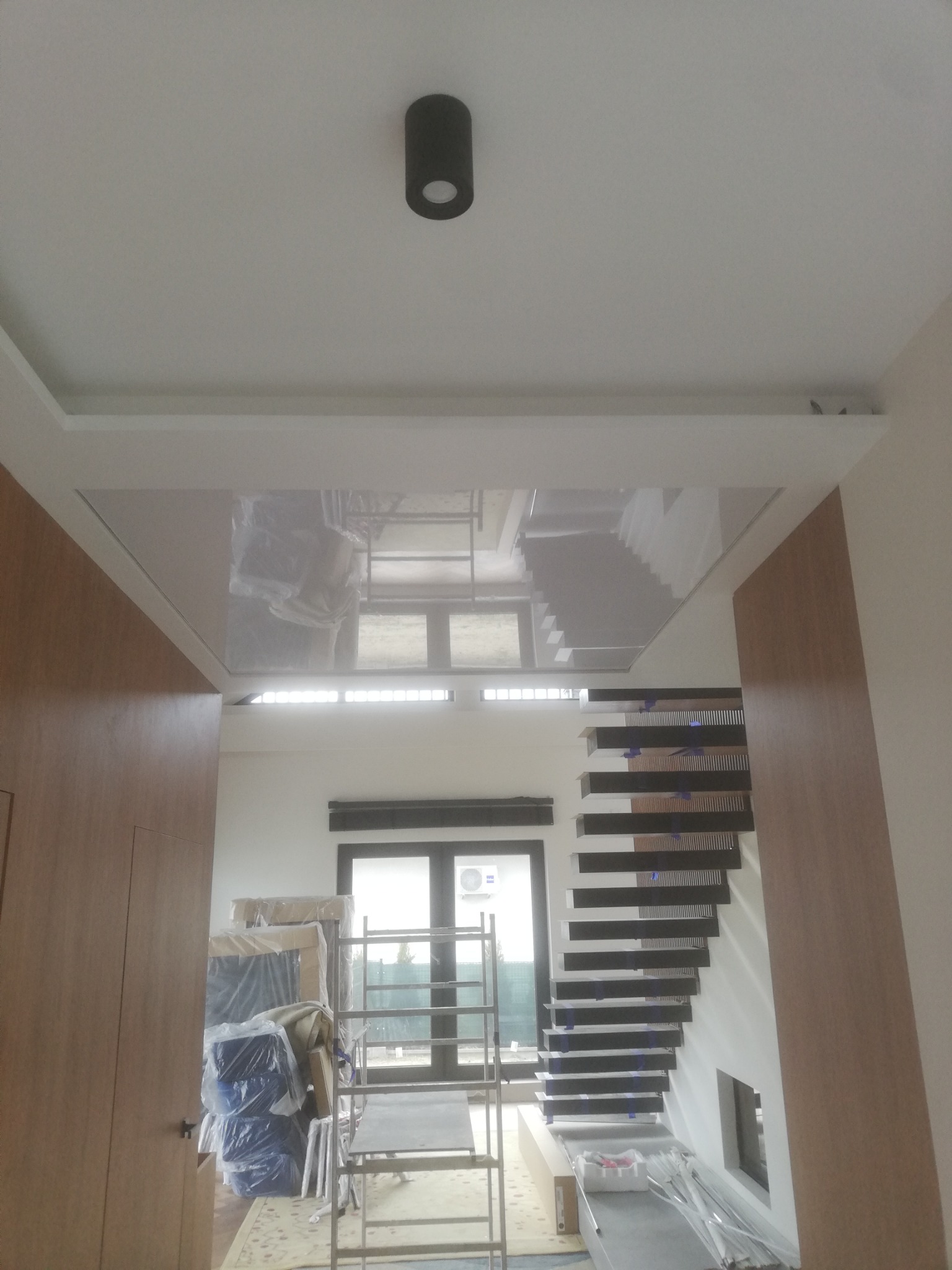 Biały sufit napinany z odbiciem schodów i okna, czarna lampa sufitowa, widoczne prace wykończeniowe i zabezpieczone meble folią.