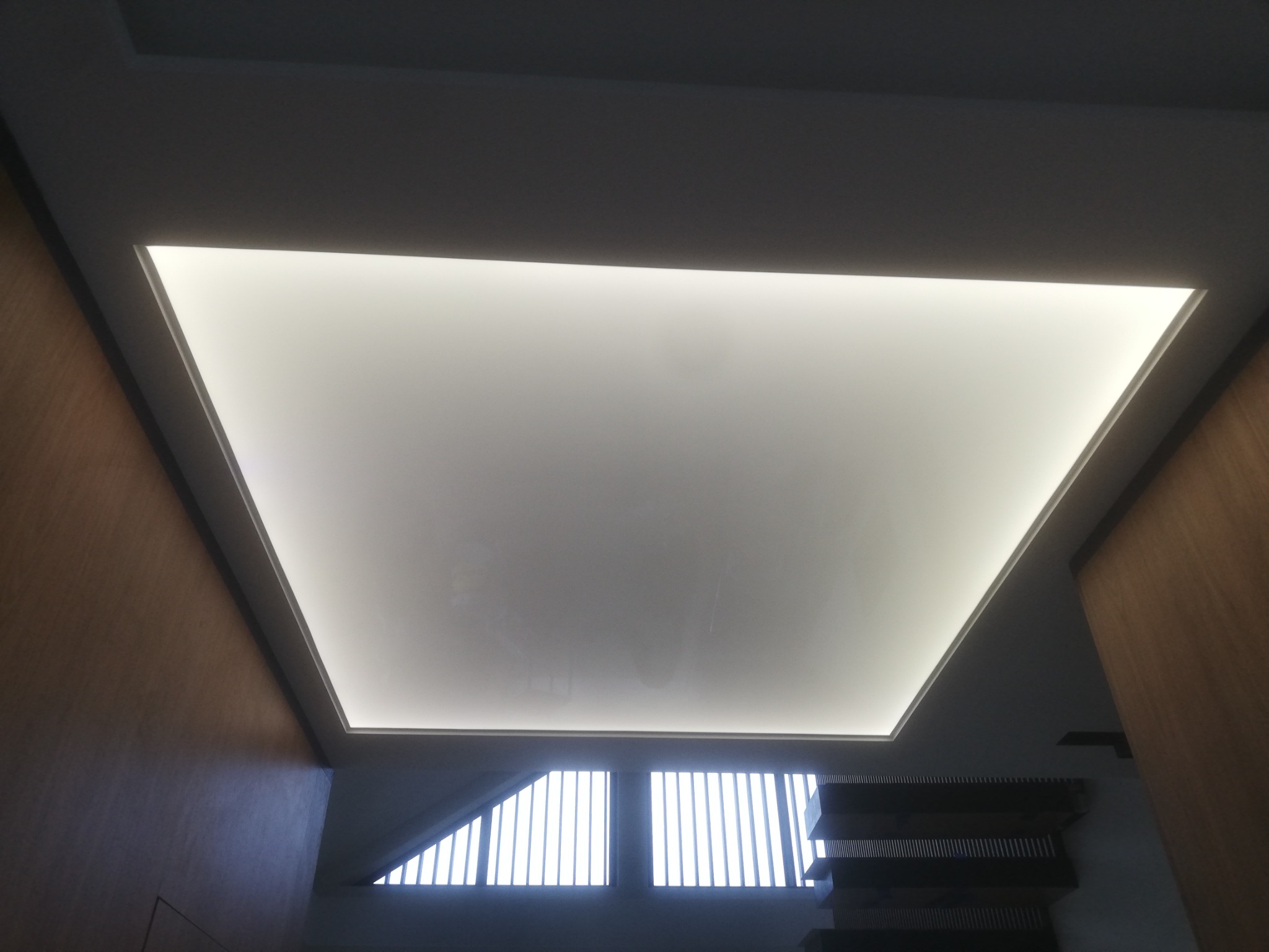 Prostokątny sufit napinany z podświetleniem LED, widok z dołu, drewniane panele ścienne po bokach, nowoczesne wnętrze.