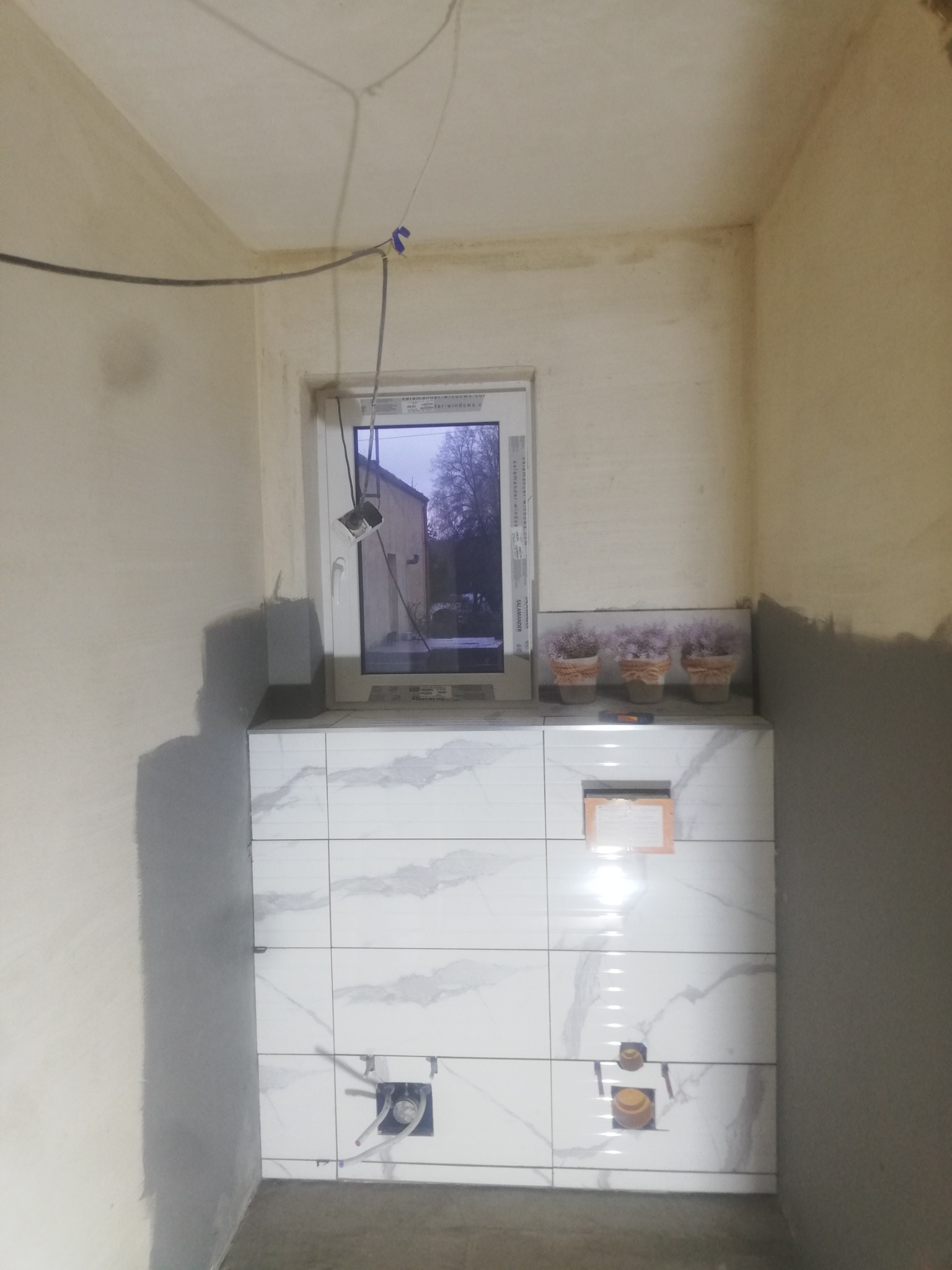 Wnętrze łazienki w trakcie prac wykończeniowych; widoczne białe płytki imitujące marmur, surowe ściany w szarym tynku, wystające rury instalacji wodnej i okno z widokiem na zewnątrz, trzy małe...