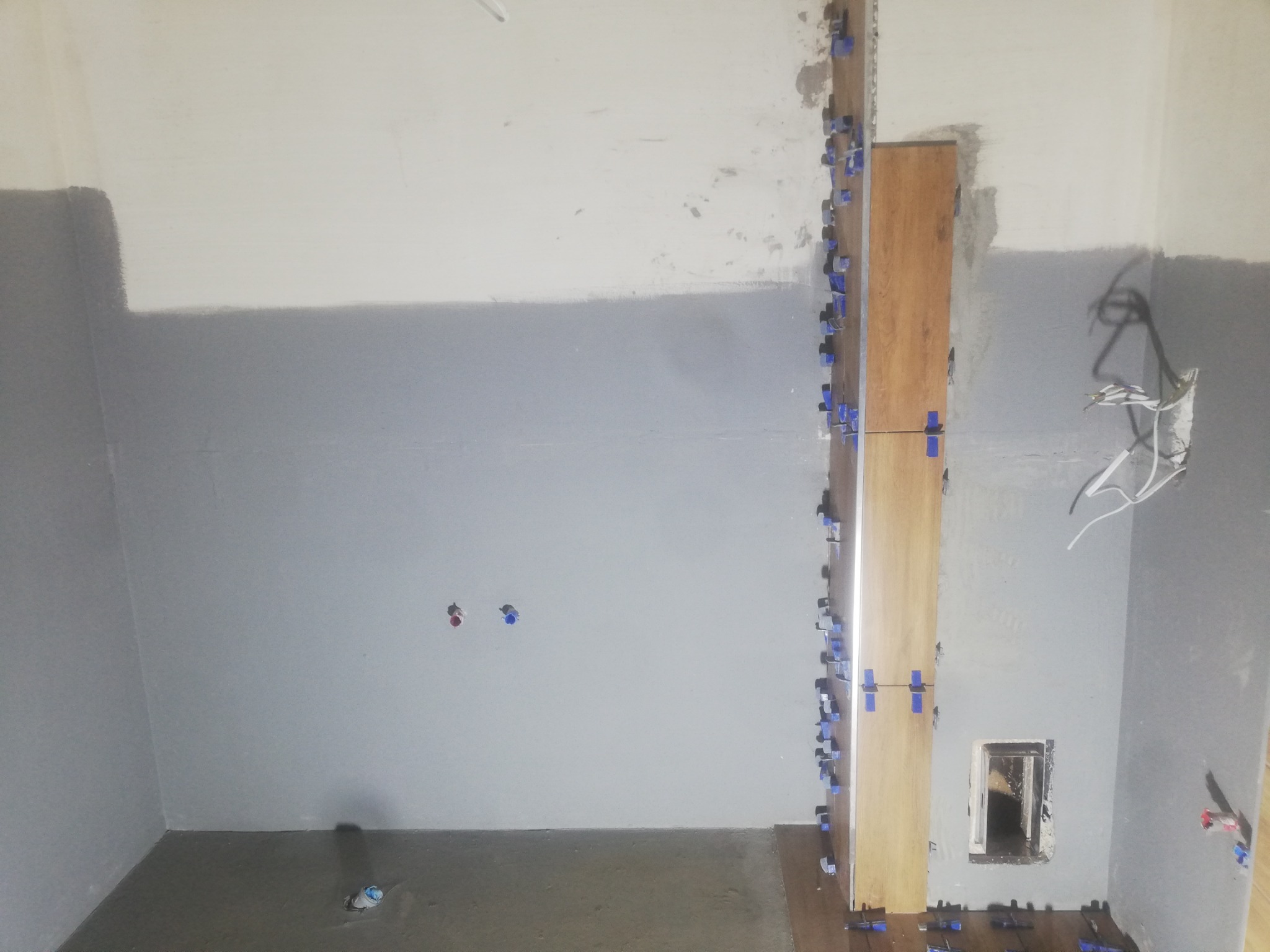 Ściana łazienki w trakcie remontu: częściowo pomalowana na szaro, z zamontowanymi pionowo płytkami imitującymi drewno przy użyciu niebieskich klipsów poziomujących, widoczne wystające kable i otwór...
