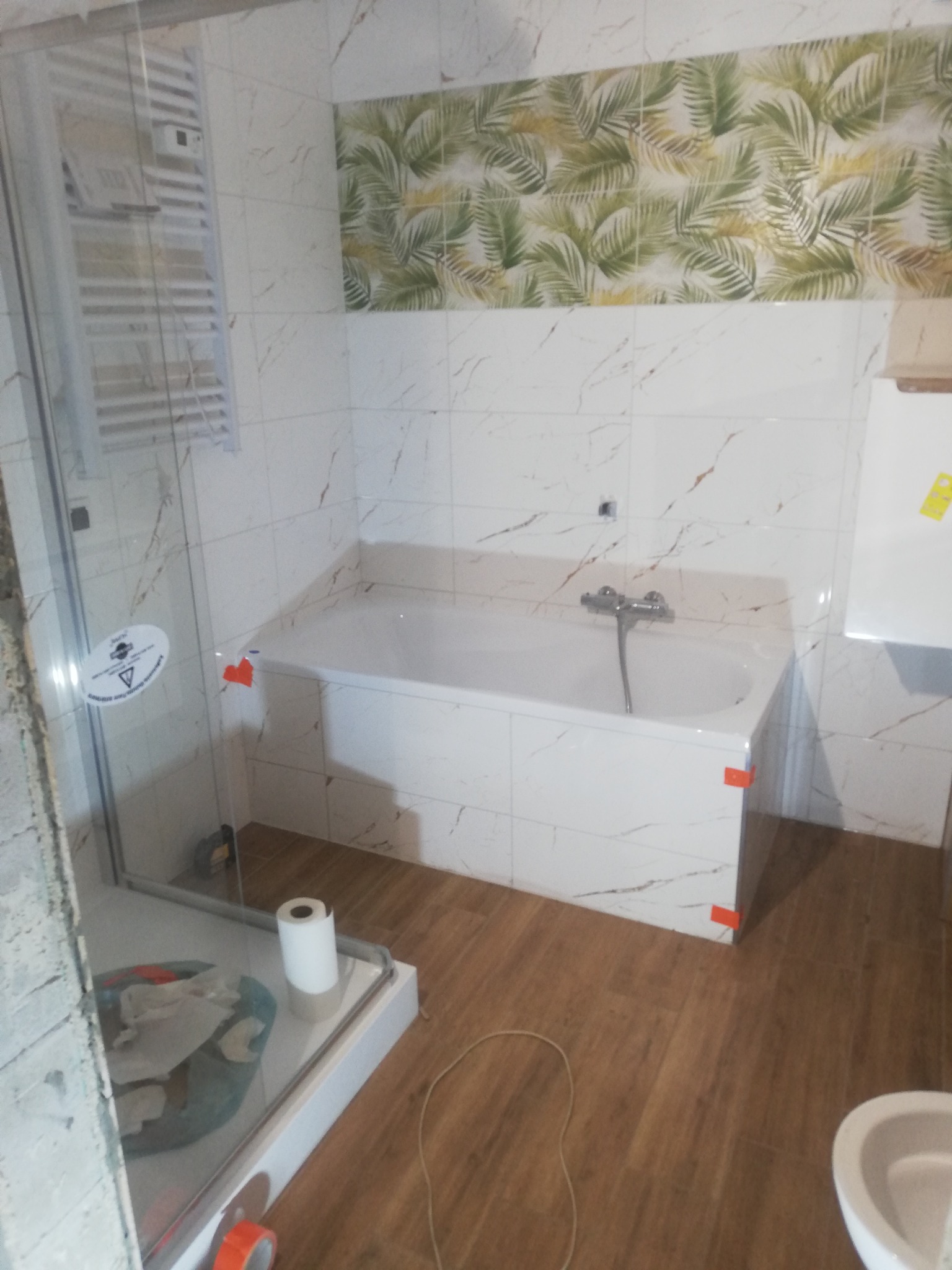 Nowoczesna łazienka z wanną obudowaną płytkami imitującymi marmur, kabiną prysznicową ze szklanymi drzwiami, drewnopodobną podłogą i akcentem w postaci płytek z motywem liści palmowych nad wanną.