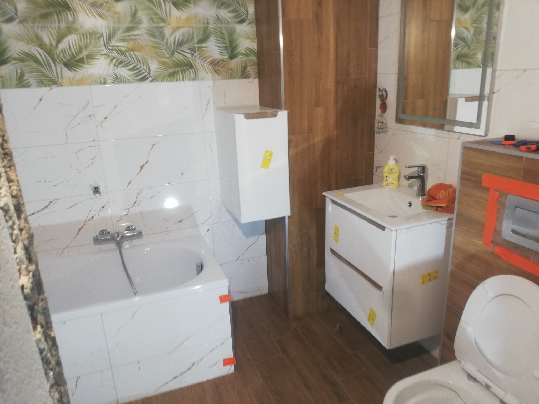 Wykończona łazienka z wanną obudowaną białymi płytkami imitującymi marmur, szafką pod umywalką i wiszącą szafką, brązowe płytki na podłodze i ścianie, fragment toalety w dolnym rogu.