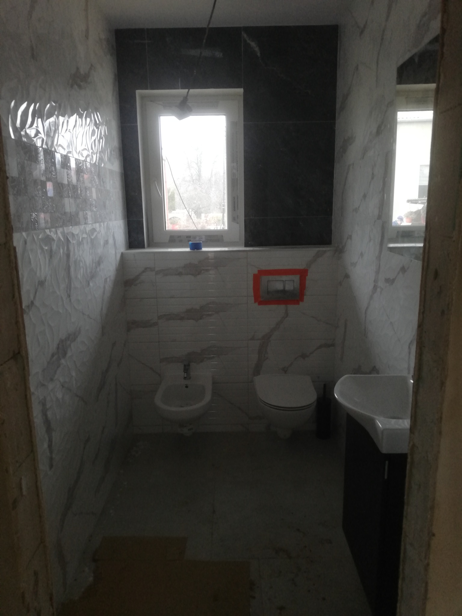 Wnętrze łazienki w trakcie remontu, widoczne białe i czarne płytki imitujące marmur na ścianach, zamontowana toaleta, bidet i umywalka z szafką, fragment okna z widokiem na zewnątrz, niedokończone...