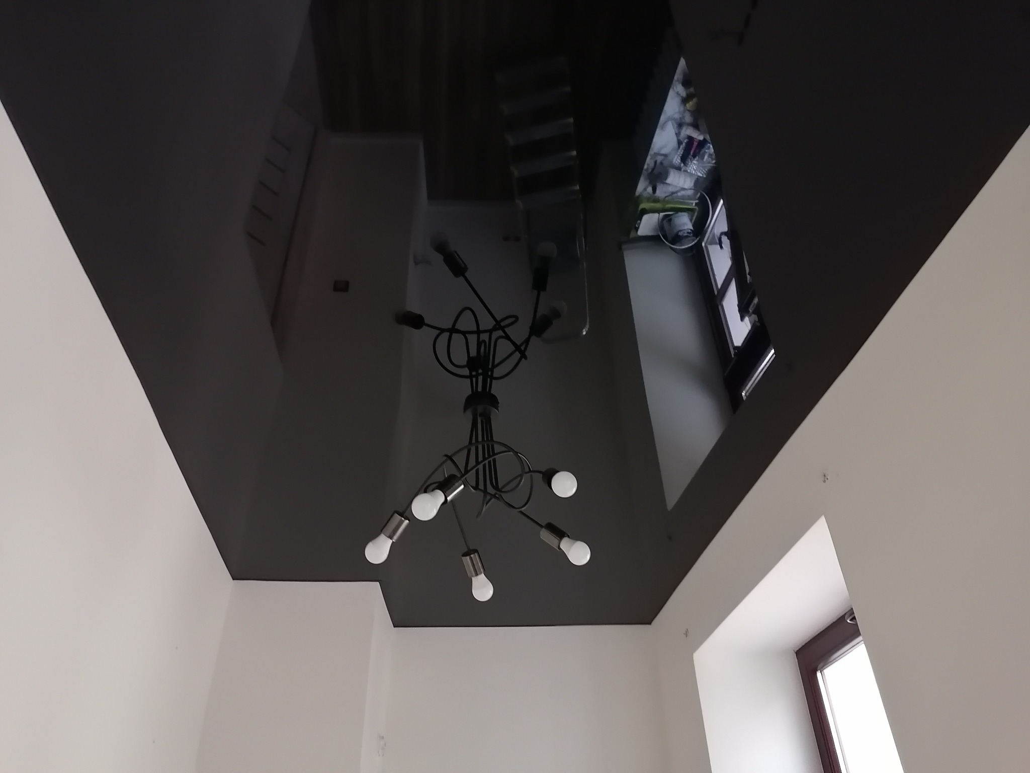 Czarny, błyszczący sufit napinany z odbiciem pomieszczenia i nowoczesną lampą wiszącą z sześcioma żarówkami w pokoju z białymi ścianami i oknem.