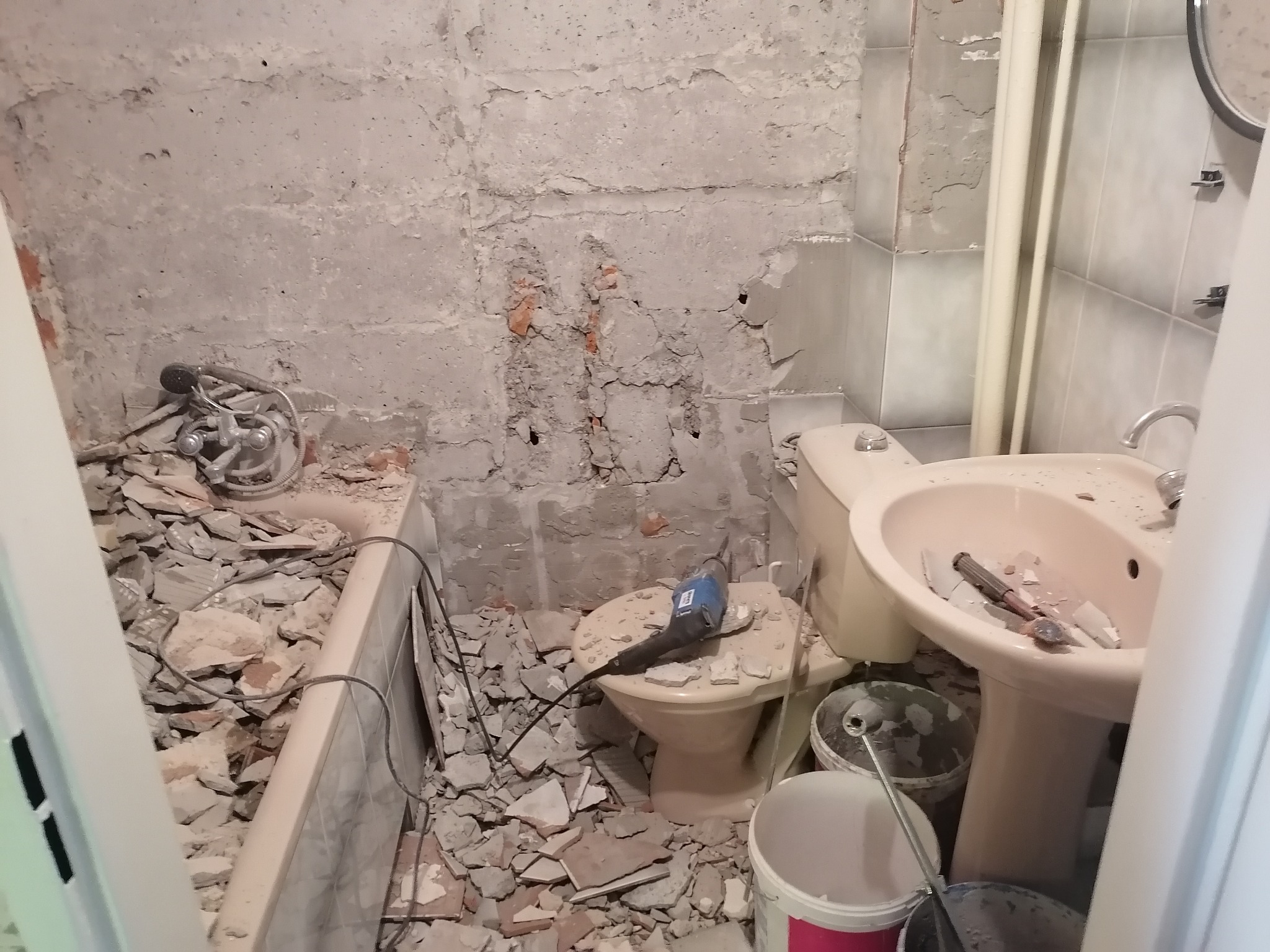 Łazienka w trakcie demontażu: gruz na podłodze i w wannie, odsłonięte ściany z resztkami płytek, wiertarka na sedesie, umywalka na postumencie i wiadra z farbą.