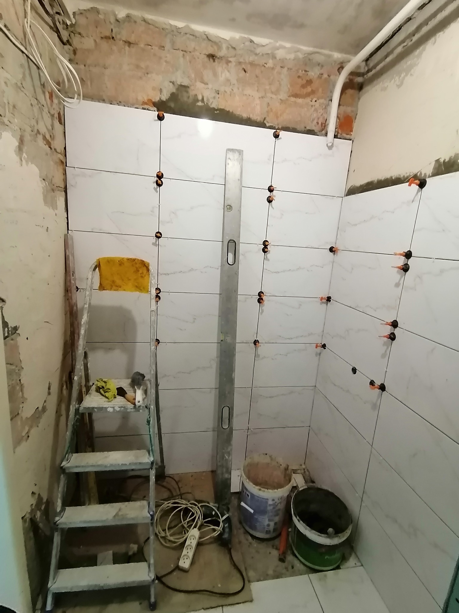 Białe płytki imitujące marmur układane na ścianie łazienki, z użyciem systemu poziomowania płytek, widoczna drabina, poziomica i wiaderka z zaprawą.