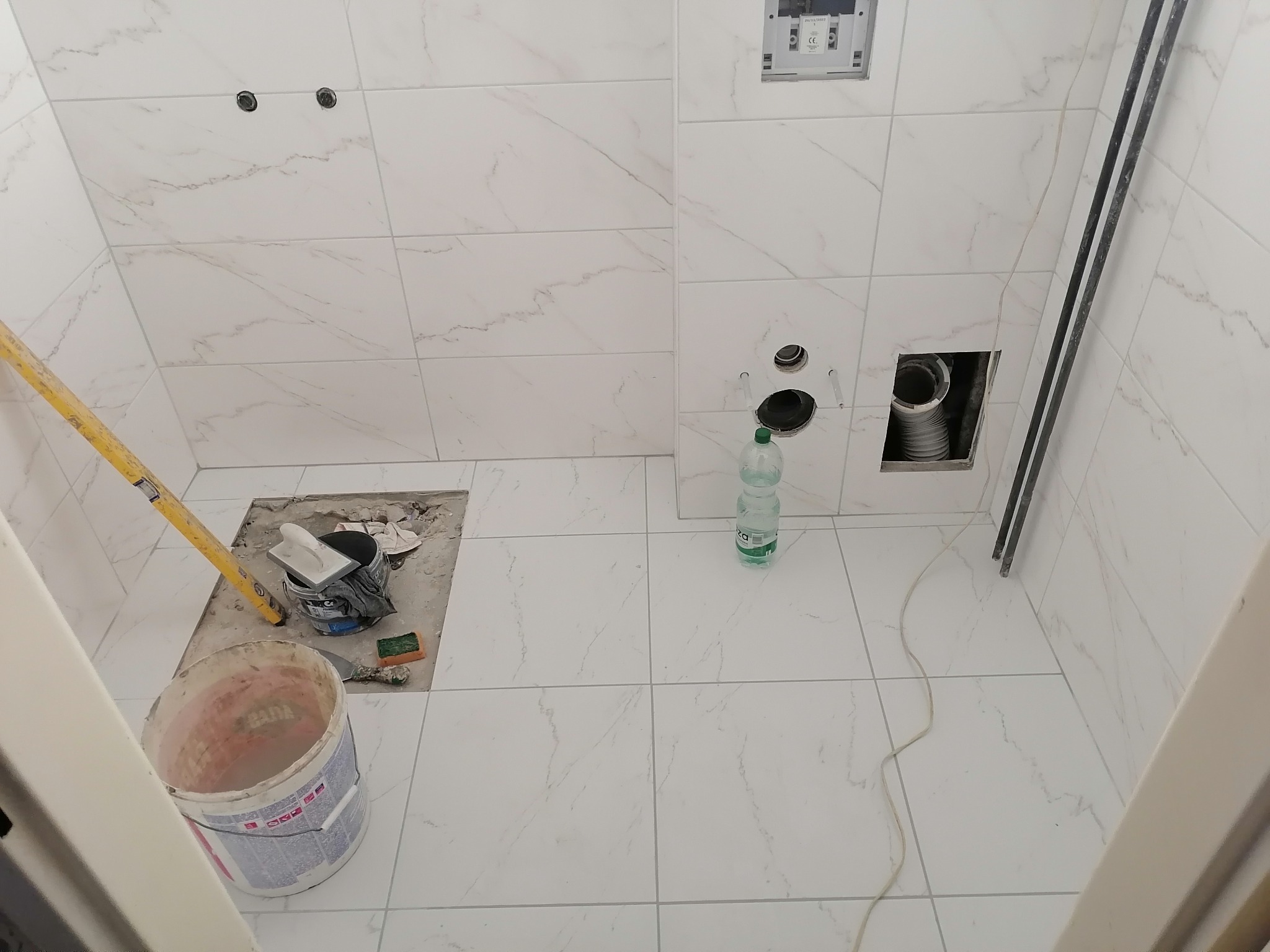 Układanie białych płytek imitujących marmur w łazience w Lublinie, widoczne narzędzia, poziomica, wiadro z klejem, otwory na instalacje sanitarne w ścianie.