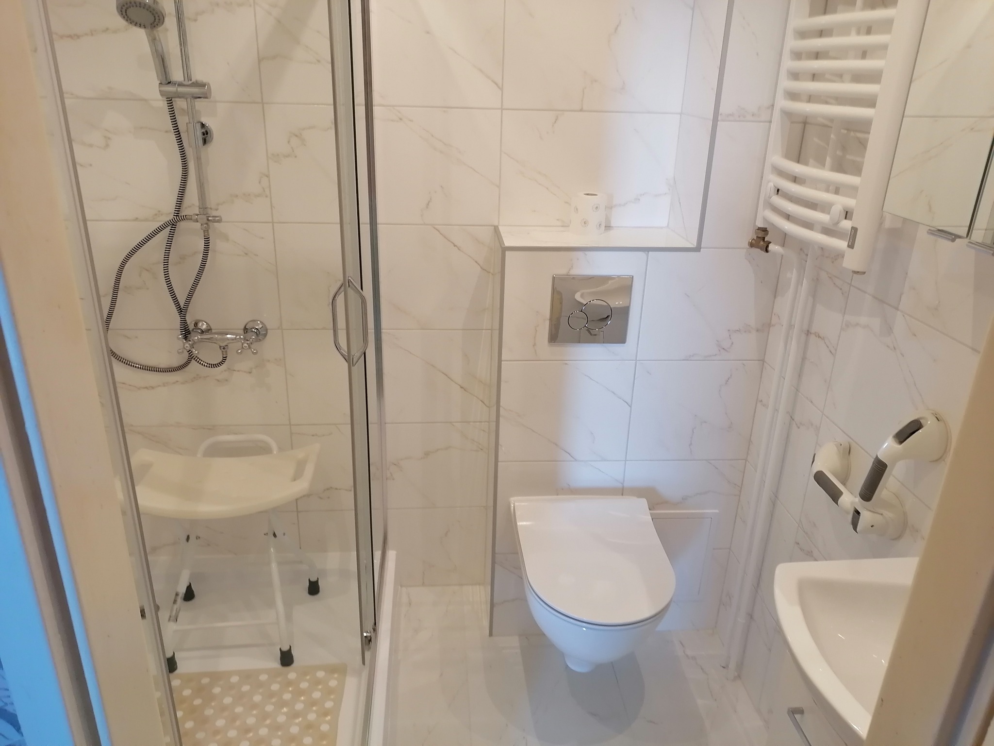 Jasna łazienka po remoncie z kabiną prysznicową, sedesem, umywalką z uchwytami dla niepełnosprawnych i grzejnikiem drabinkowym na tle białych płytek imitujących marmur.