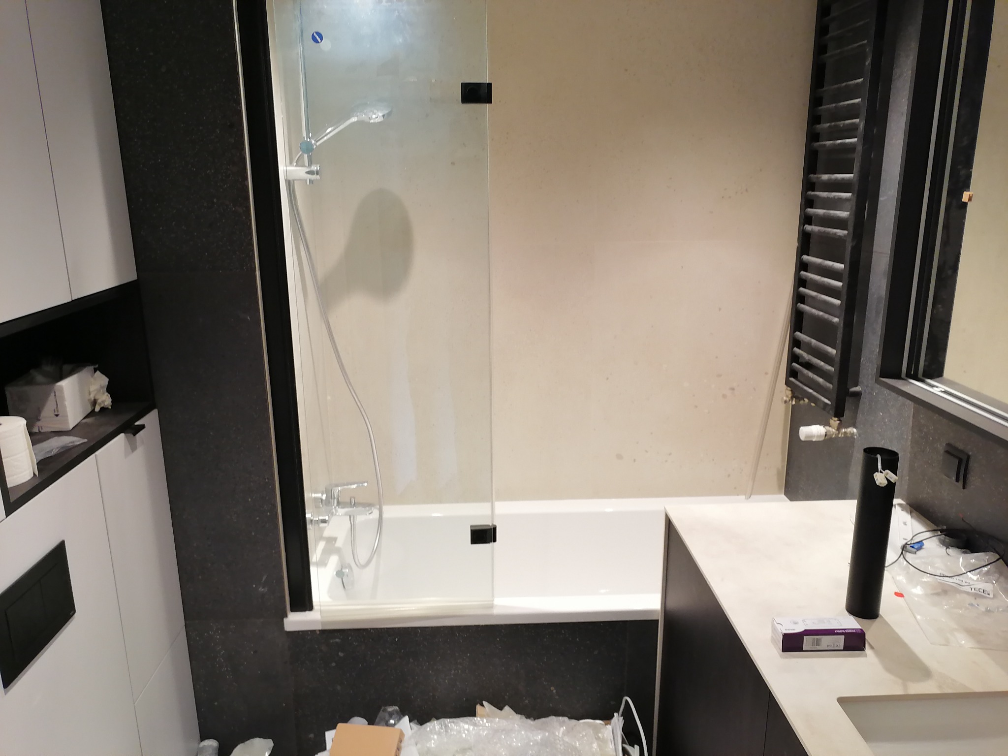 Nowoczesna łazienka z białą wanną za szklanym parawanem, czarne płytki na ścianie, grzejnik drabinkowy i białe szafki, widoczne ślady po remoncie.