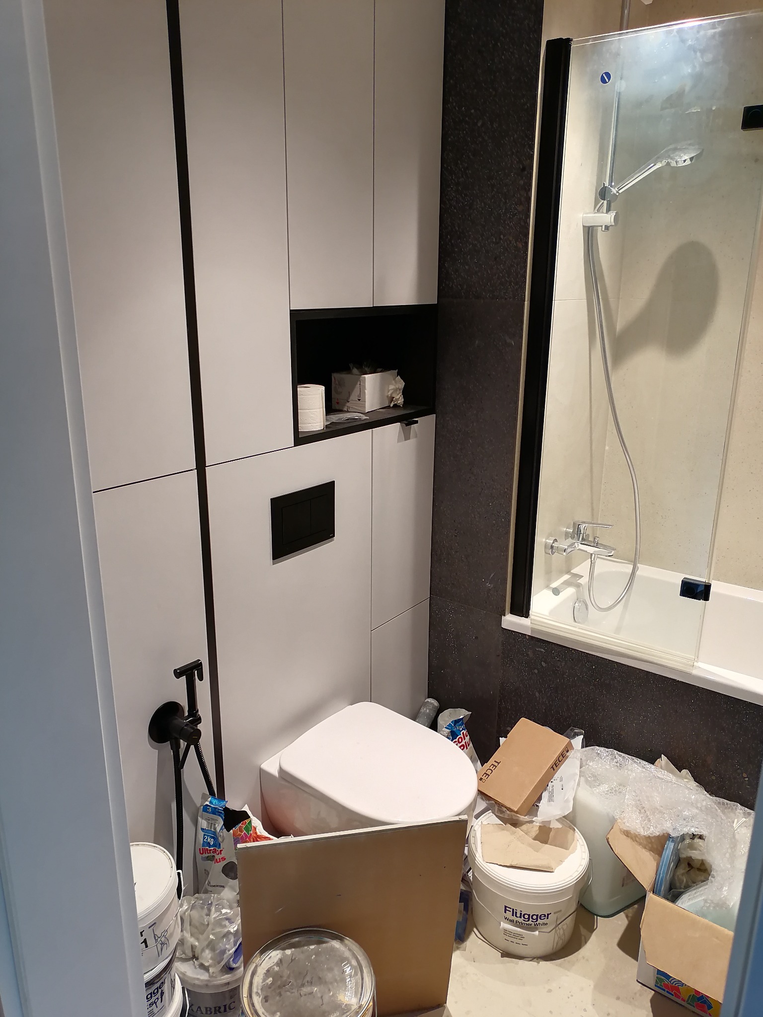 Łazienka w trakcie remontu: widoczna toaleta, wanna z prysznicem, zabudowa meblowa w kolorze białym oraz puszki z farbą i inne materiały budowlane na podłodze.