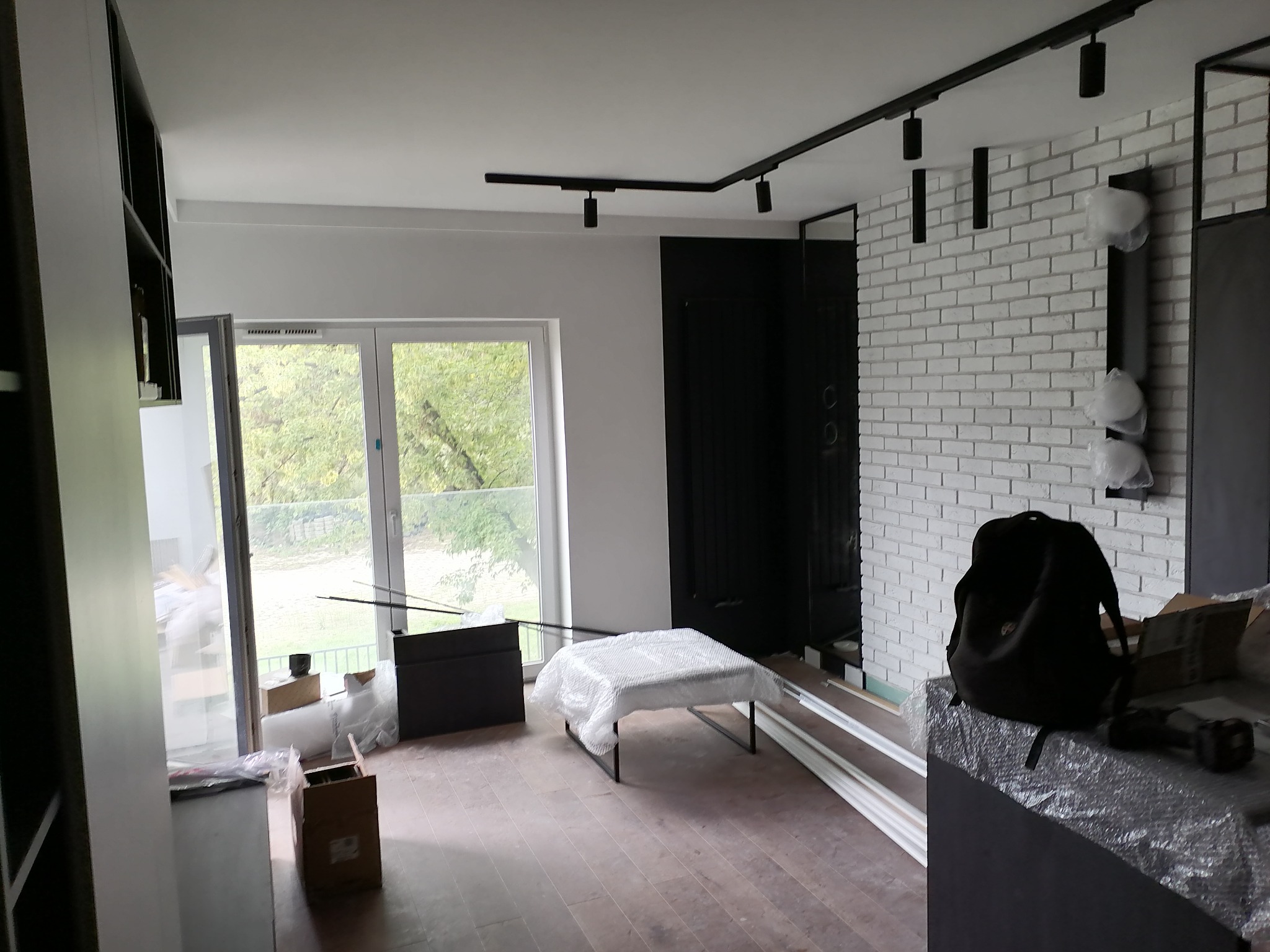 Wnętrze pokoju w trakcie remontu: biała ceglana ściana, czarne oświetlenie szynowe, podłoga z paneli, okno z widokiem na zieleń, meble zabezpieczone folią, widoczny plecak i narzędzia.