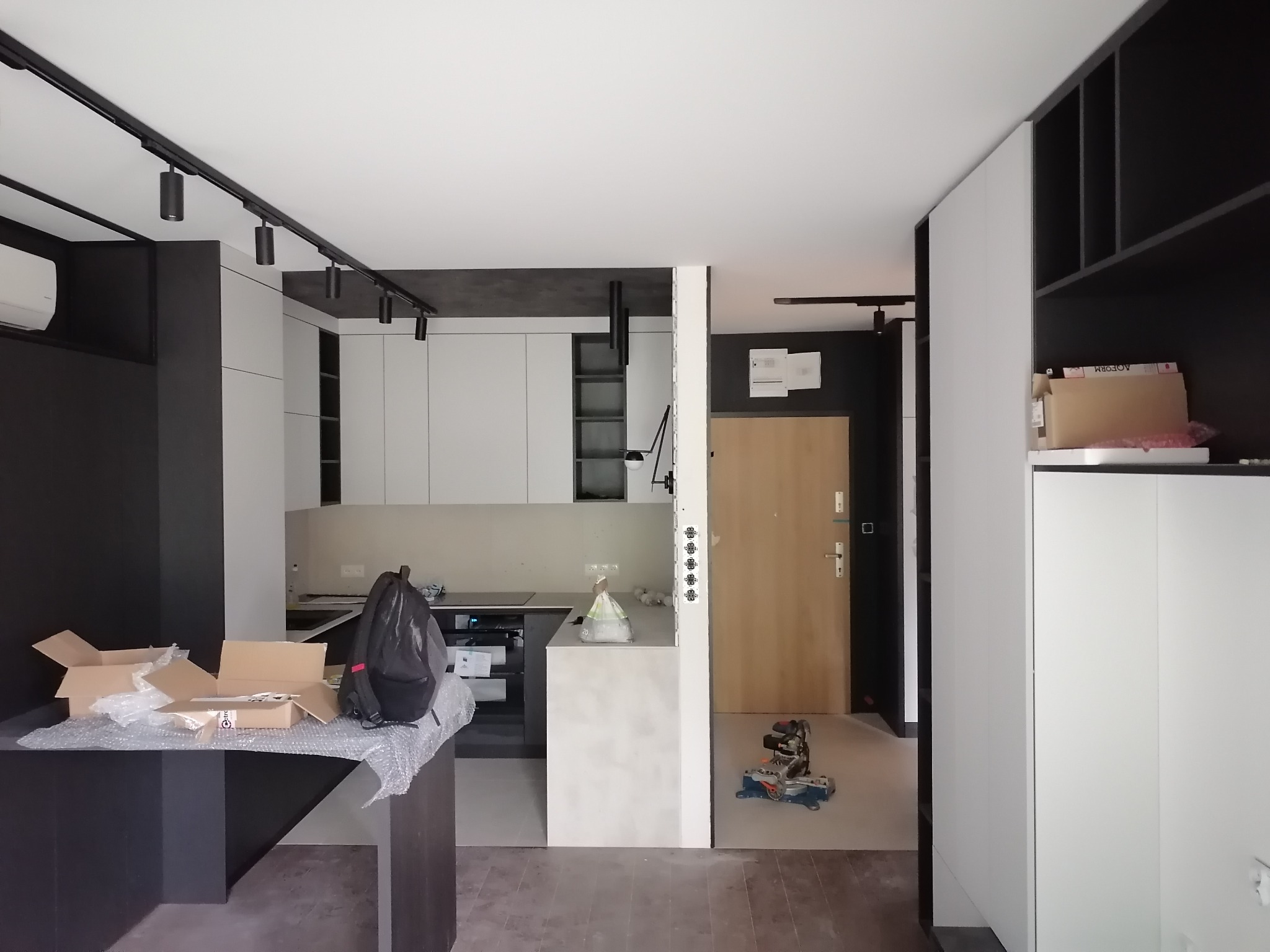 Wnętrze mieszkania w trakcie wykończenia: widoczne białe szafki kuchenne, czarne elementy dekoracyjne, rozpakowane kartony na stole, torba i piła ukośnica na podłodze.