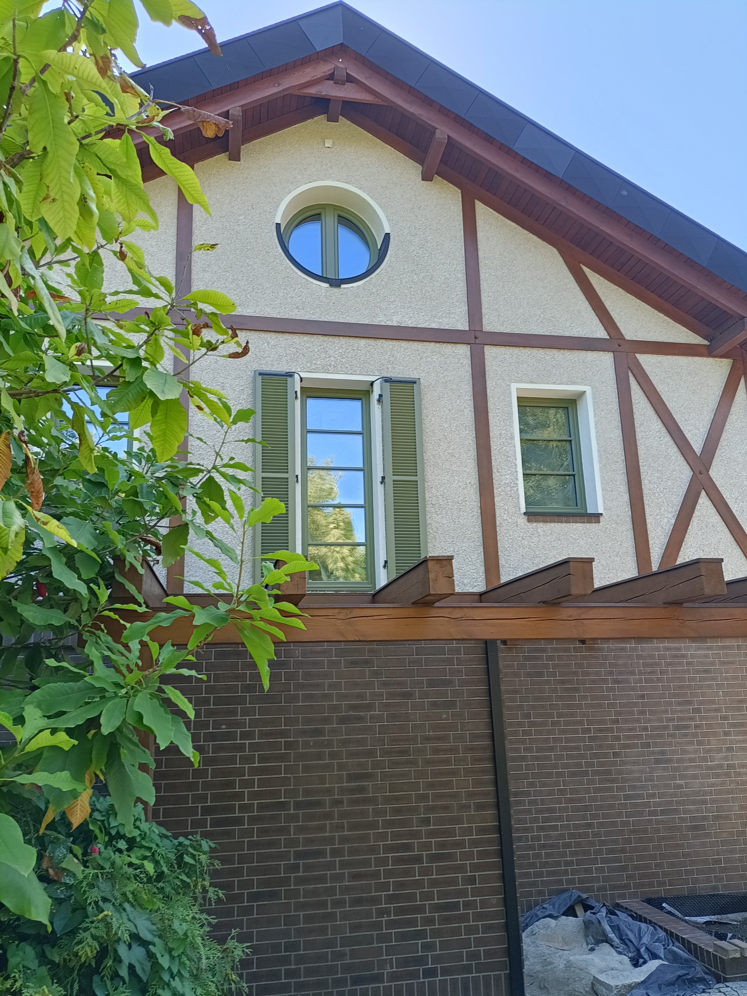 Elewacja domu z elementami szachulca, okrągłym oknem i zielonymi okiennicami, częściowo zasłonięta zielenią.