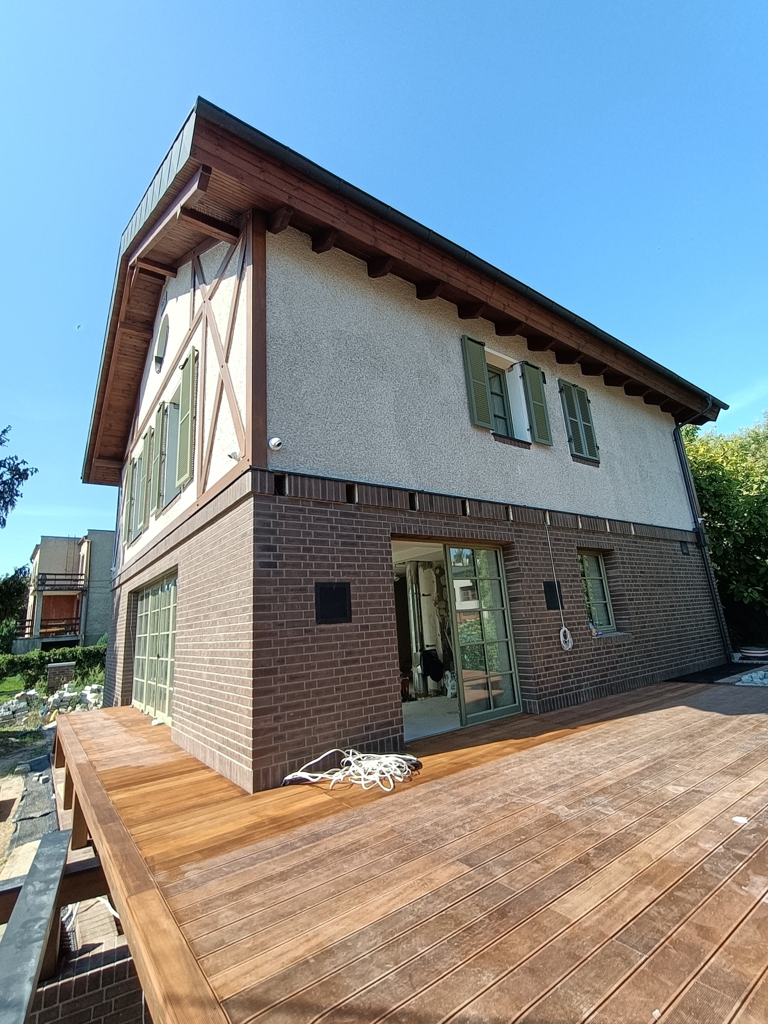 Dwukondygnacyjny dom z ceglaną elewacją do połowy wysokości, drewnianą konstrukcją szkieletową na górnej kondygnacji, zielonymi okiennicami i nowym drewnianym tarasem z widocznym białym sznurem.