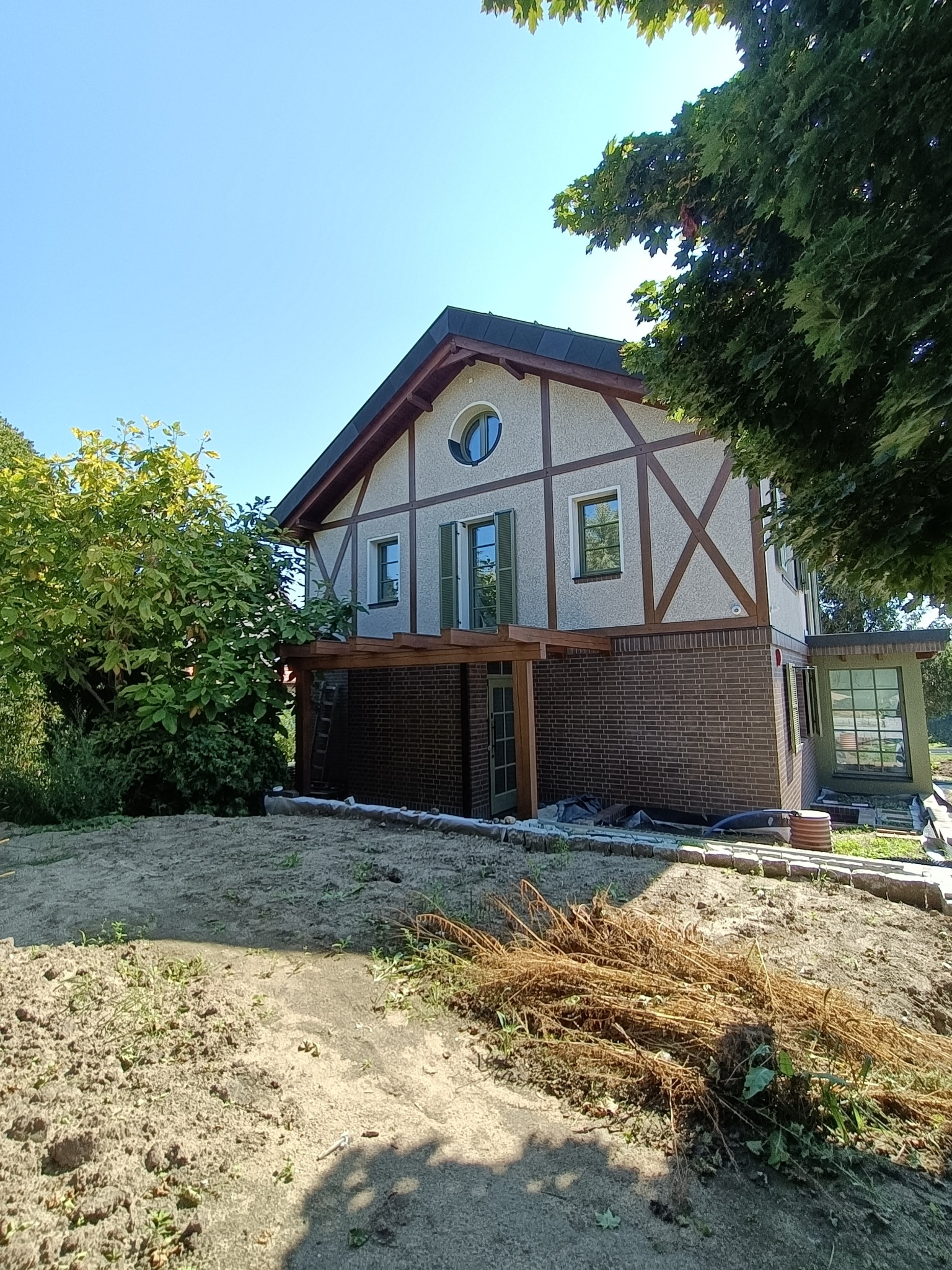 Dwupiętrowy dom z ceglaną podstawą i jasną elewacją w stylu szachulcowym, z drewnianą pergolą i zielonymi okiennicami, widziany w słoneczny dzień z perspektywy ogrodu w trakcie prac wykończeniowych.