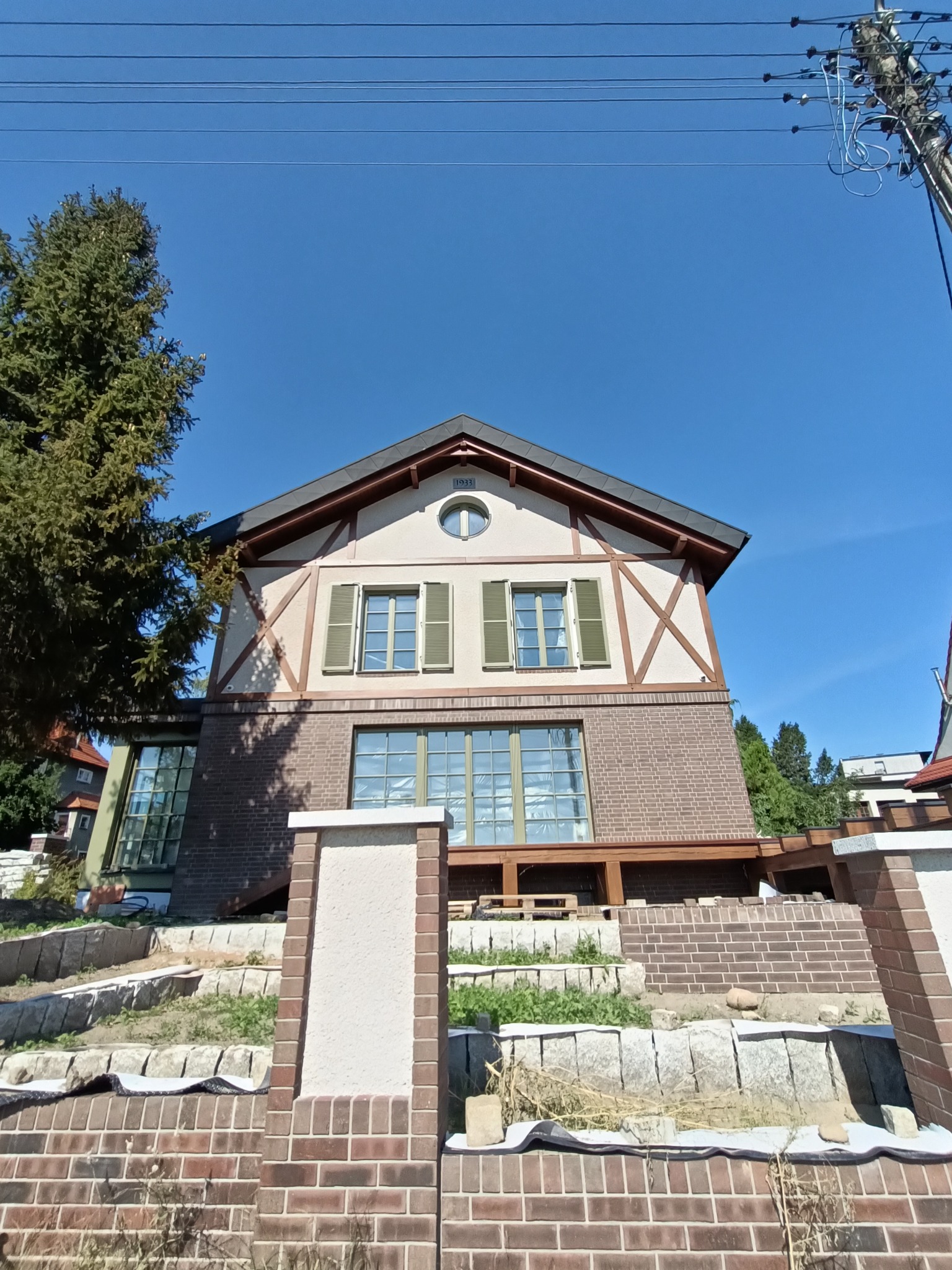 Dwupiętrowy dom z elementami muru pruskiego, ceglaną elewacją na parterze i zielonymi okiennicami, widziany z dołu, z częściowo ukończonym murem oporowym z cegły na pierwszym planie.