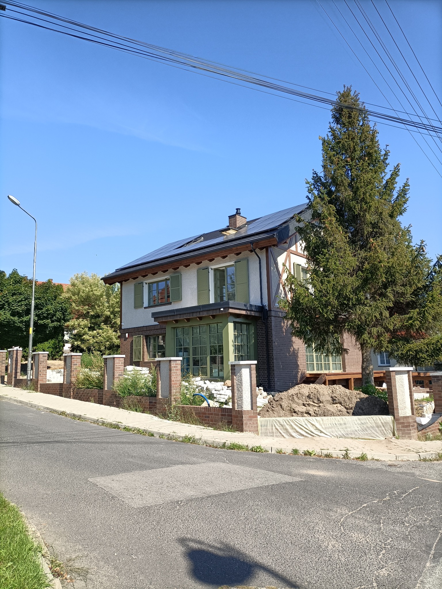 Dwukondygnacyjny dom z panelami słonecznymi na dachu i częściowo wykończonym ceglanym ogrodzeniem od frontu, z widocznymi śladami prac budowlanych.