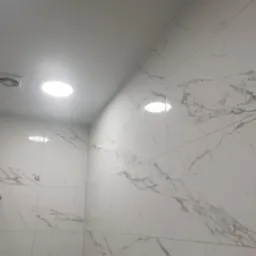 Wykończona ściana łazienki z białych płytek imitujących marmur, oświetlona okrągłymi lampami sufitowymi i wyposażona w chromowany uchwyt na słuchawkę prysznicową oraz kratkę wentylacyjną.