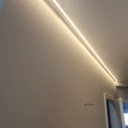 Nowoczesne oświetlenie LED wzdłuż sufitu, świecące ciepłym światłem, na tle gładkiej, szarej ściany. Widoczne fragmenty drzwi i instalacji elektrycznej.