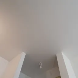 Biały, gładki sufit w trakcie wykańczania, widoczna żarówka na kablu, świeżo pomalowane ściany z widocznymi narożnikami.