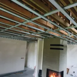 Sufit z metalową konstrukcją pod płyty gipsowo-kartonowe, widoczne drewniane belki stropowe, ściany w stanie surowym, kominek z palącym się ogniem w tle.