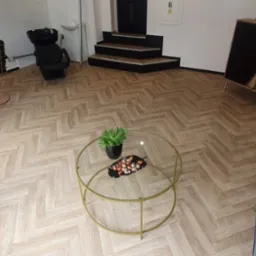 Wnętrze salonu fryzjerskiego z podłogą ułożoną w jodełkę, fotelami fryzjerskimi z miedzianymi podstawami, myjnią, stolikiem kawowym ze szklanym blatem i zieloną rośliną, oraz szafką z drewnianymi...